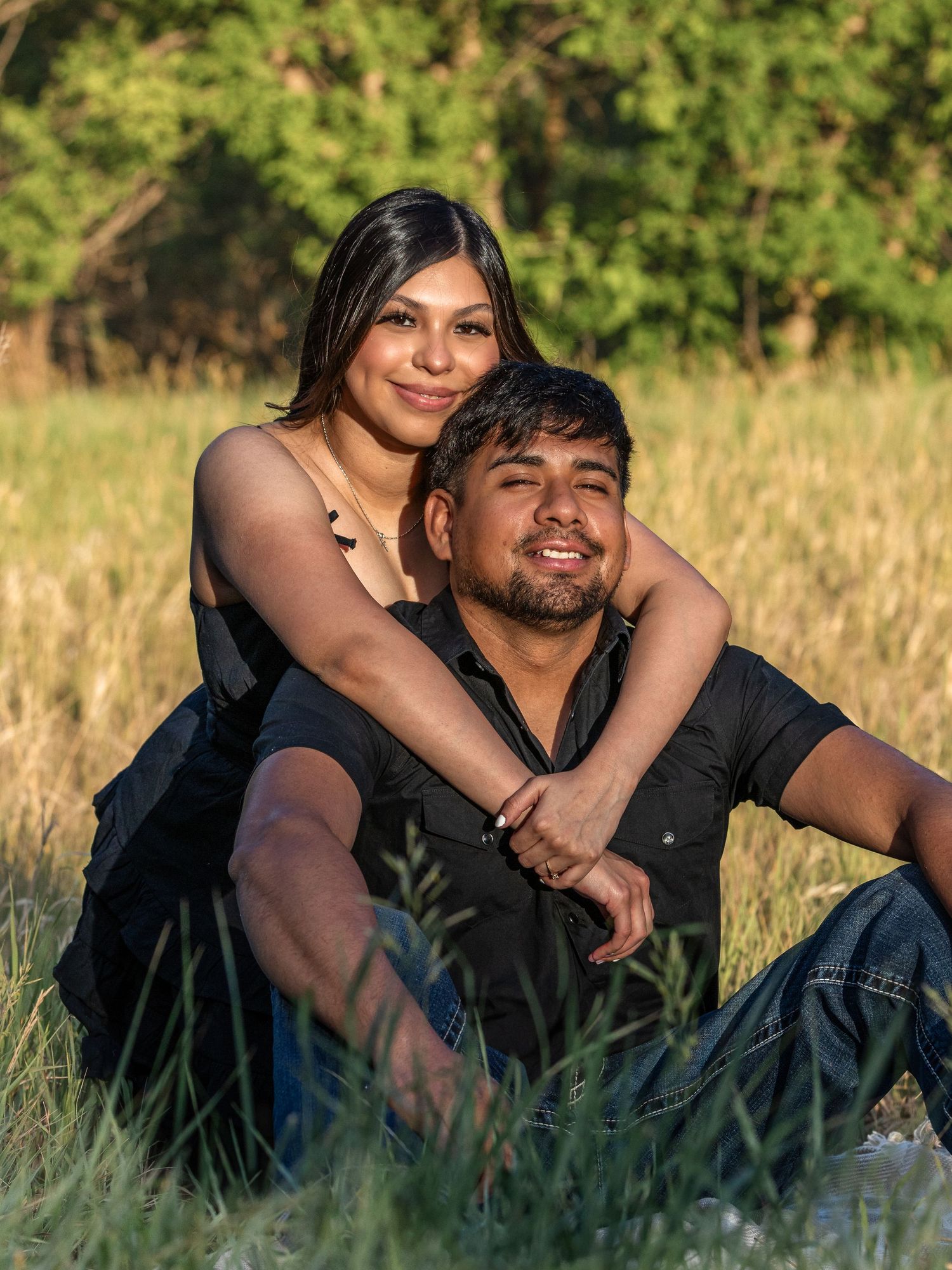 A young couple sits in a grassy field, smiling warmly, with the woman embracing the man from behind at golden hour.