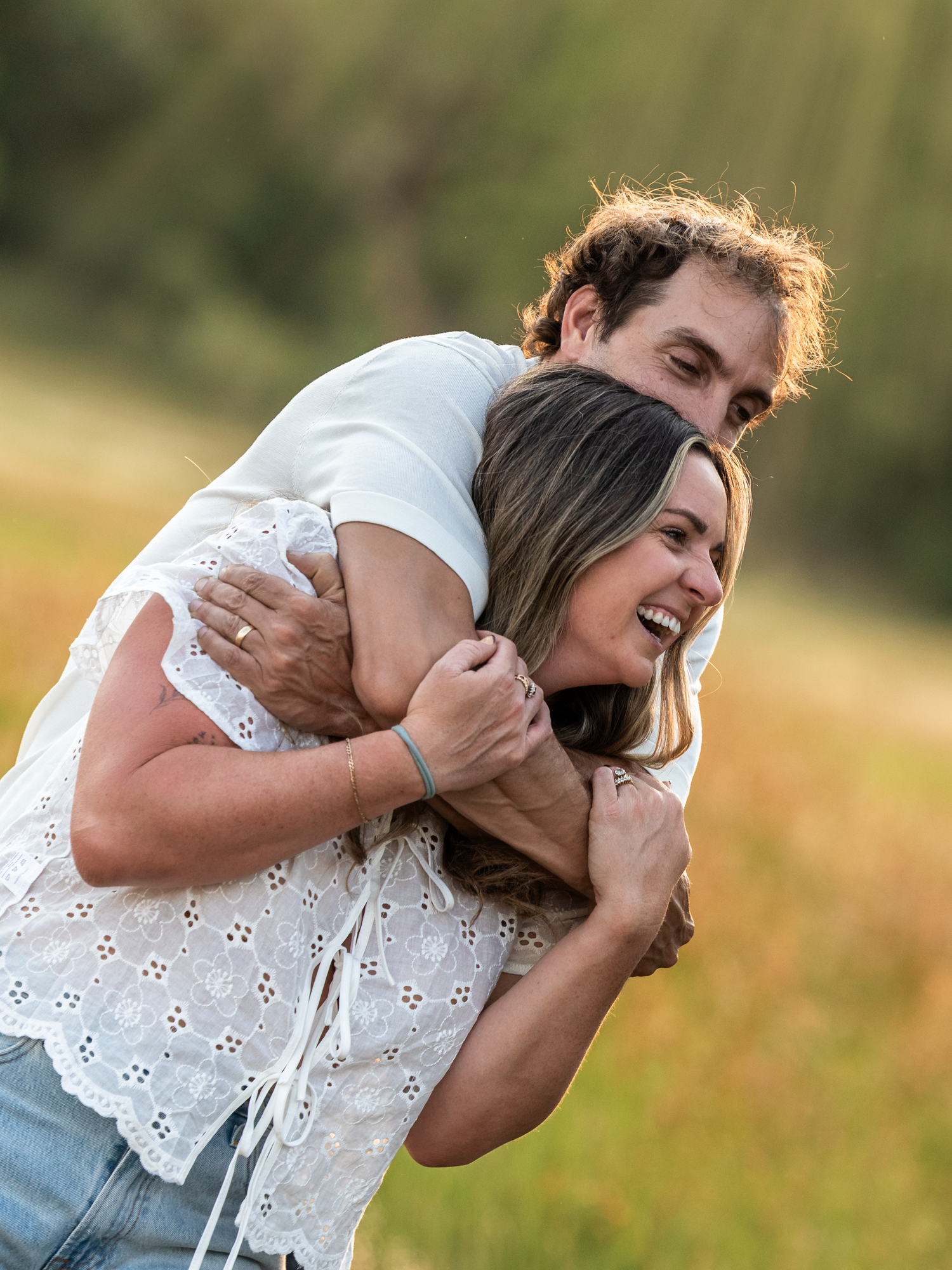 A joyful couple embraces outdoors in a sunny field, the woman laughing as the man hugs her from behind.