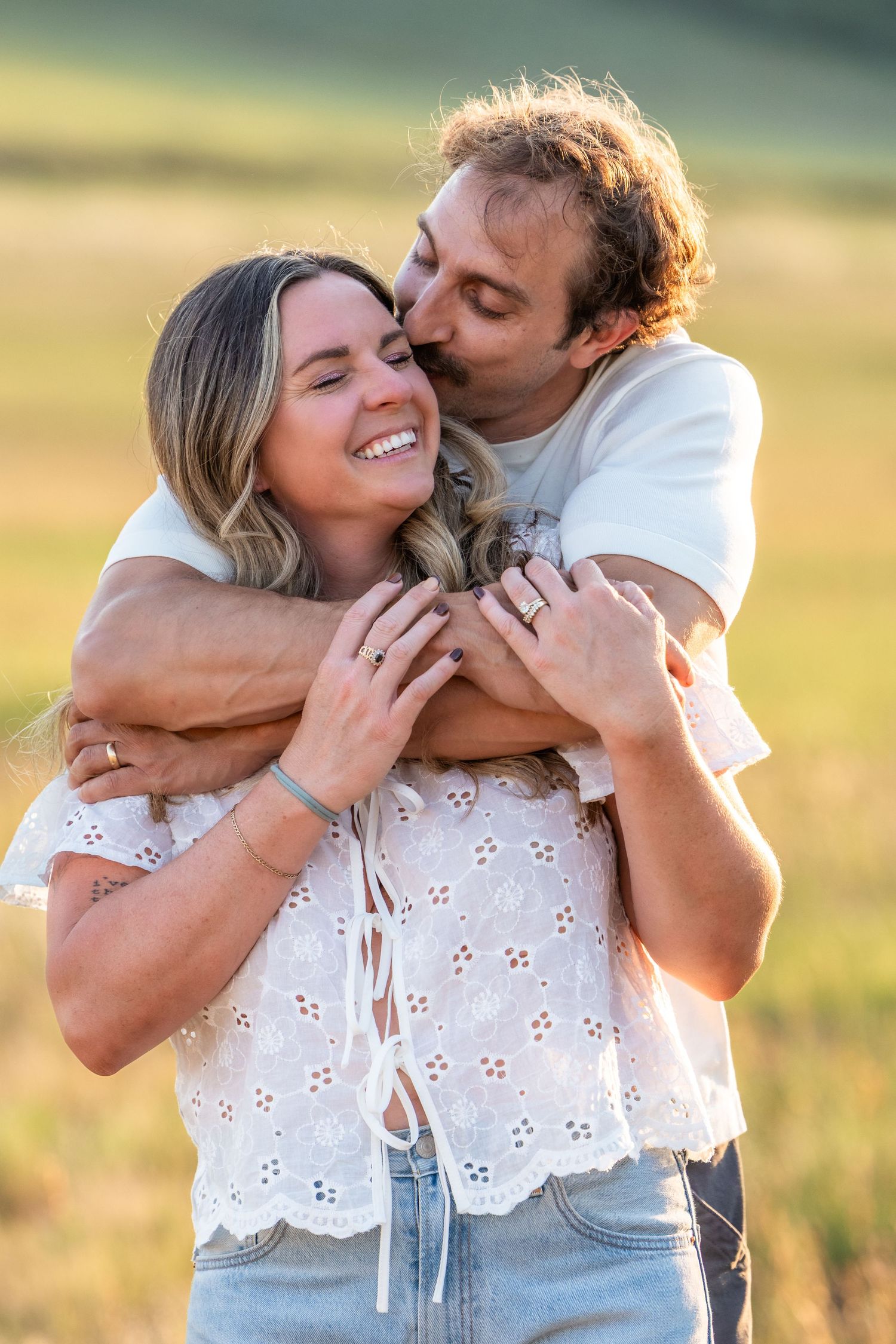 A happy couple embraces outdoors at golden hour, man kissing woman's cheek as she smiles in a white eyelet top.