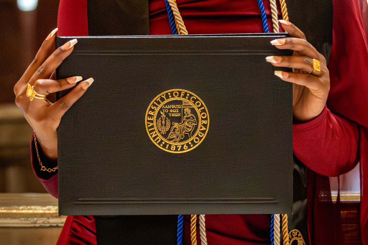 Graduate holds black diploma cover with gold university seal emblem during graduation ceremony in Littleton, Colorado.