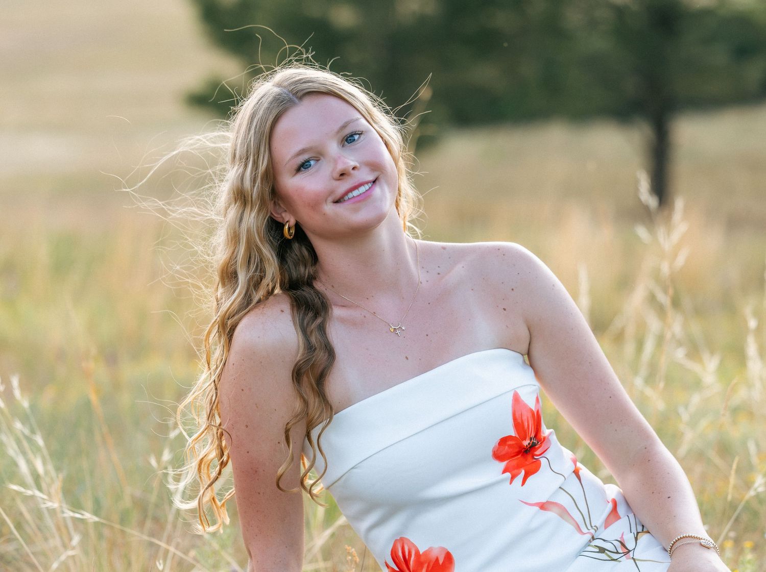 Young woman with long blonde hair in white floral dress smiling in a sunny golden field.