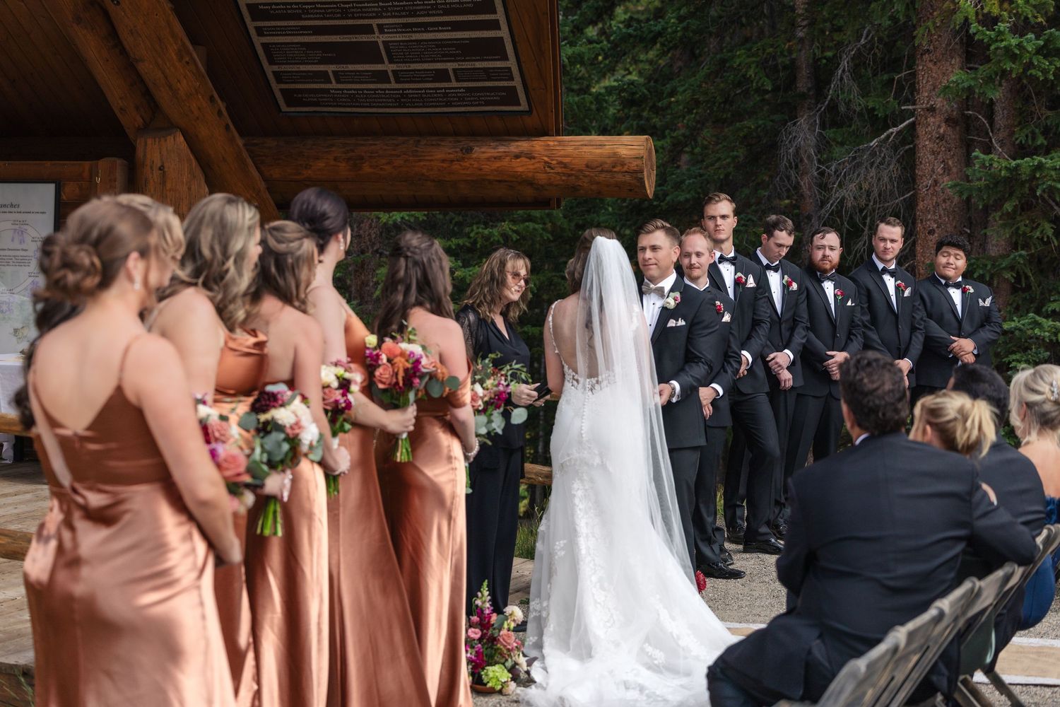 Outdoor wedding ceremony with bridesmaids in copper dresses, groomsmen in black suits, and couple at the altar in a forest setting.