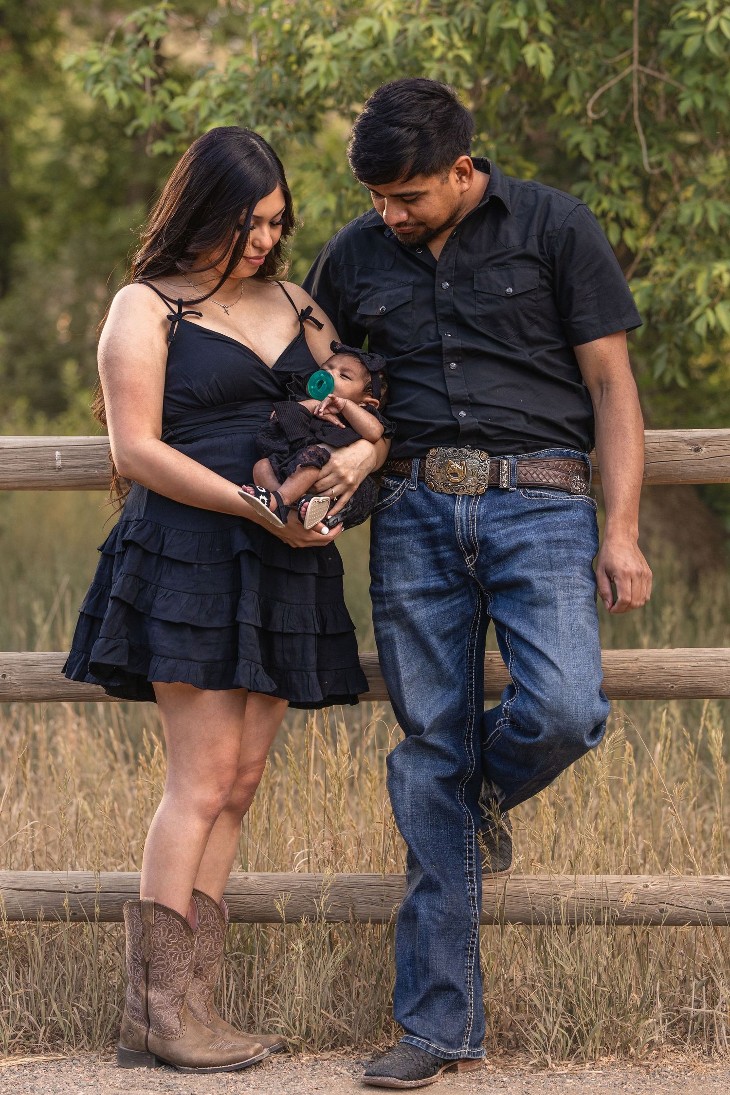 A young couple in matching black outfits and cowboy boots lovingly gaze at their newborn baby outdoors.