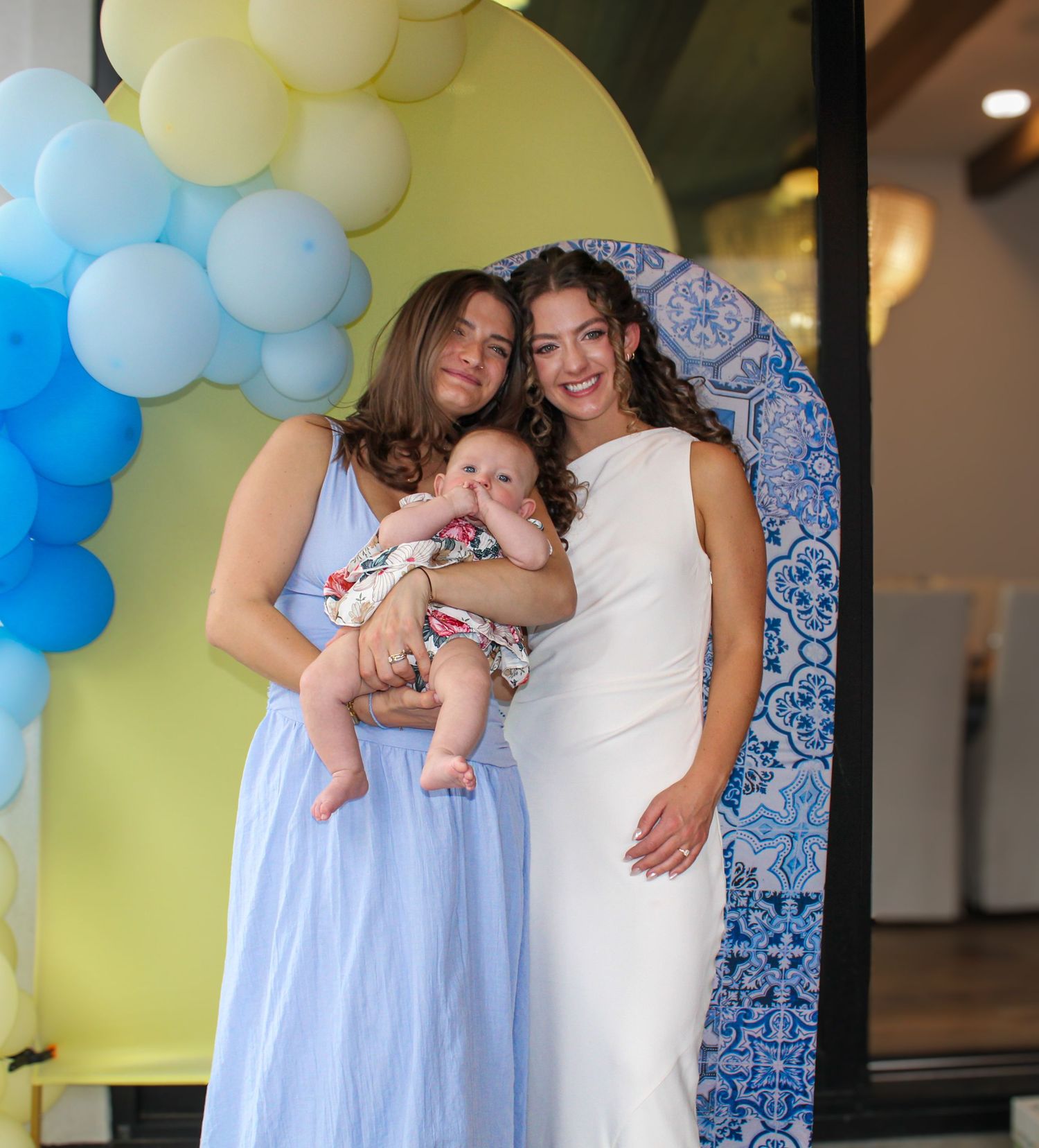 Two smiling women hold an infant baby at a festive event with colorful balloon arch decor.
