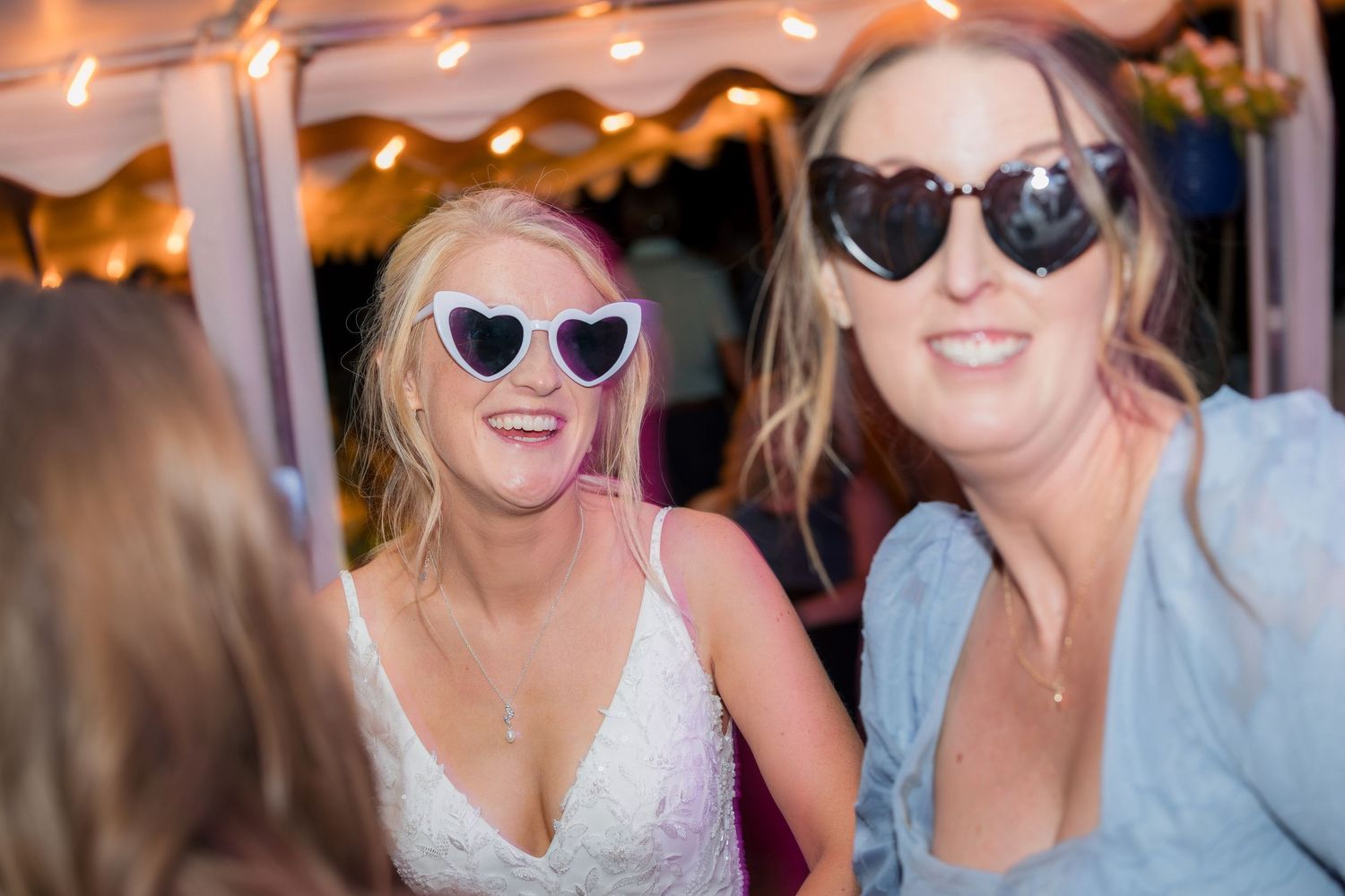 Two women wearing fun sunglasses smile and laugh together at a festive outdoor party with string lights.