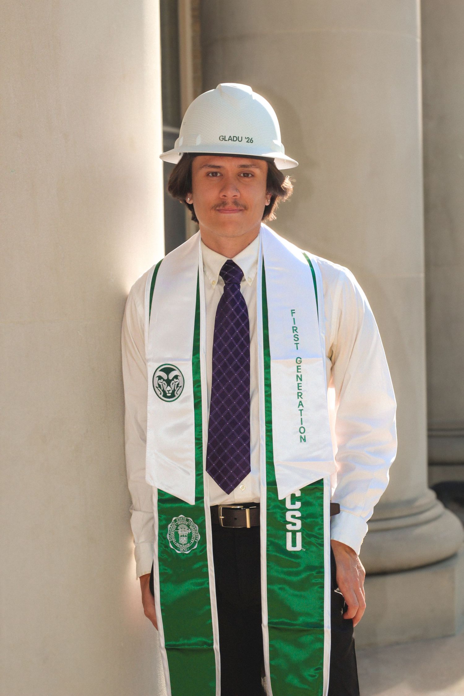 CSU graduate in white hard hat and green First Generation stole poses proudly between columns.