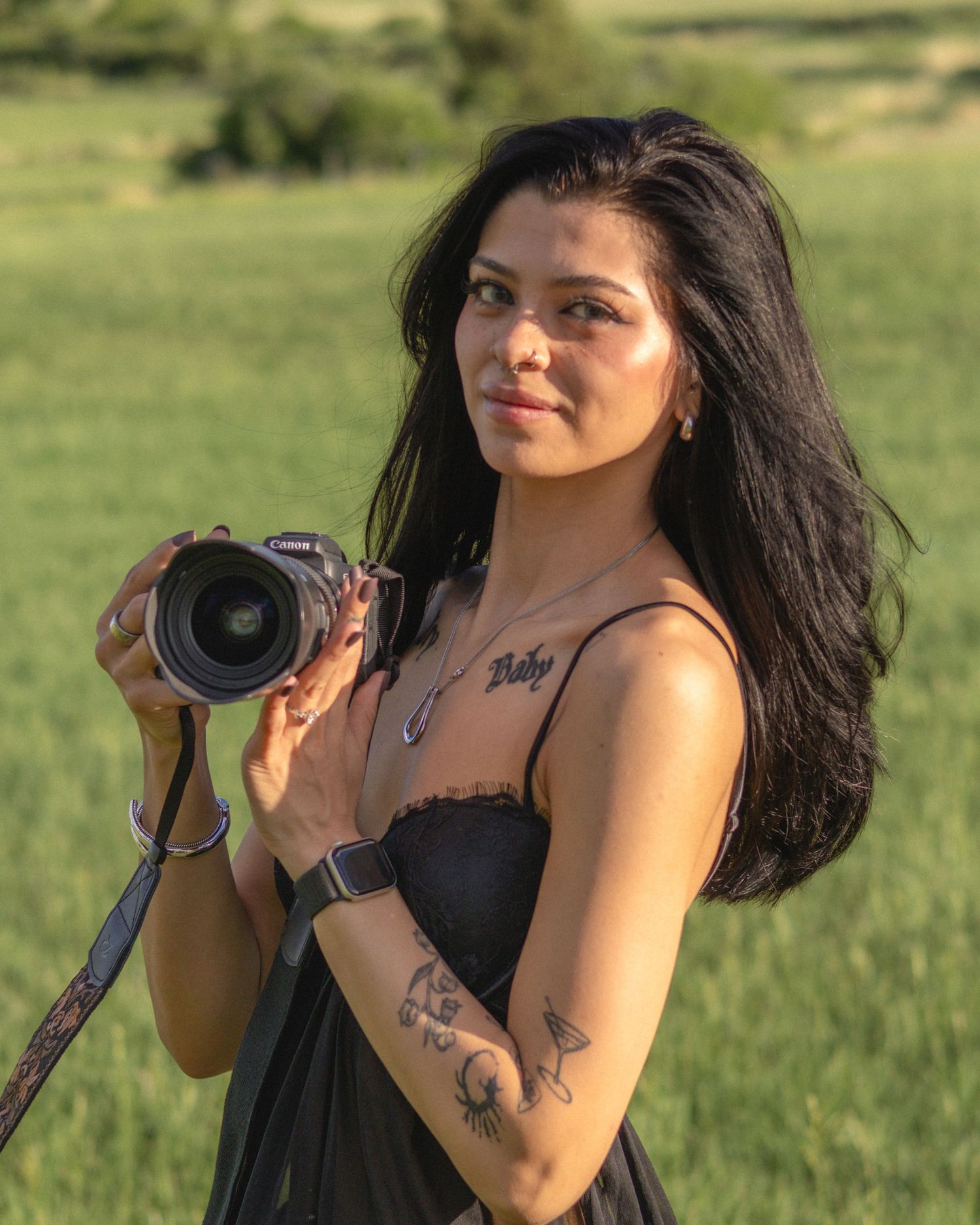 Young woman with tattoos holding a Canon camera in a green field, wearing a black dress and apple watch.