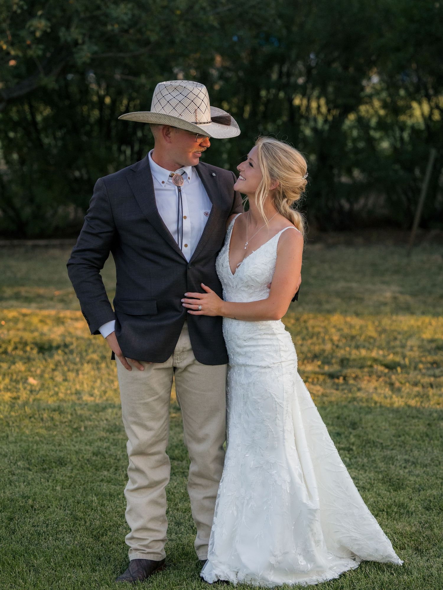 Bride in white lace gown and groom in navy blazer with cowboy hat share a tender moment on green lawn.