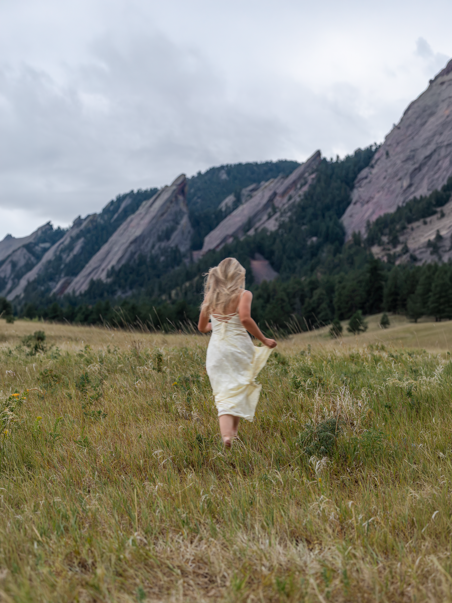 A woman in a white dress walks through a grassy meadow with the iconic Flatirons rock formations in Boulder, Colorado.