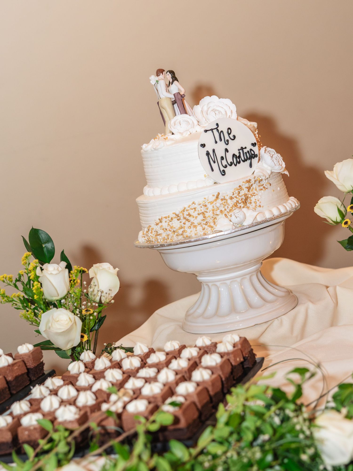 Elegant two-tier wedding cake reading The McCoys on a white pedestal stand, surrounded by white roses and chocolate desserts.