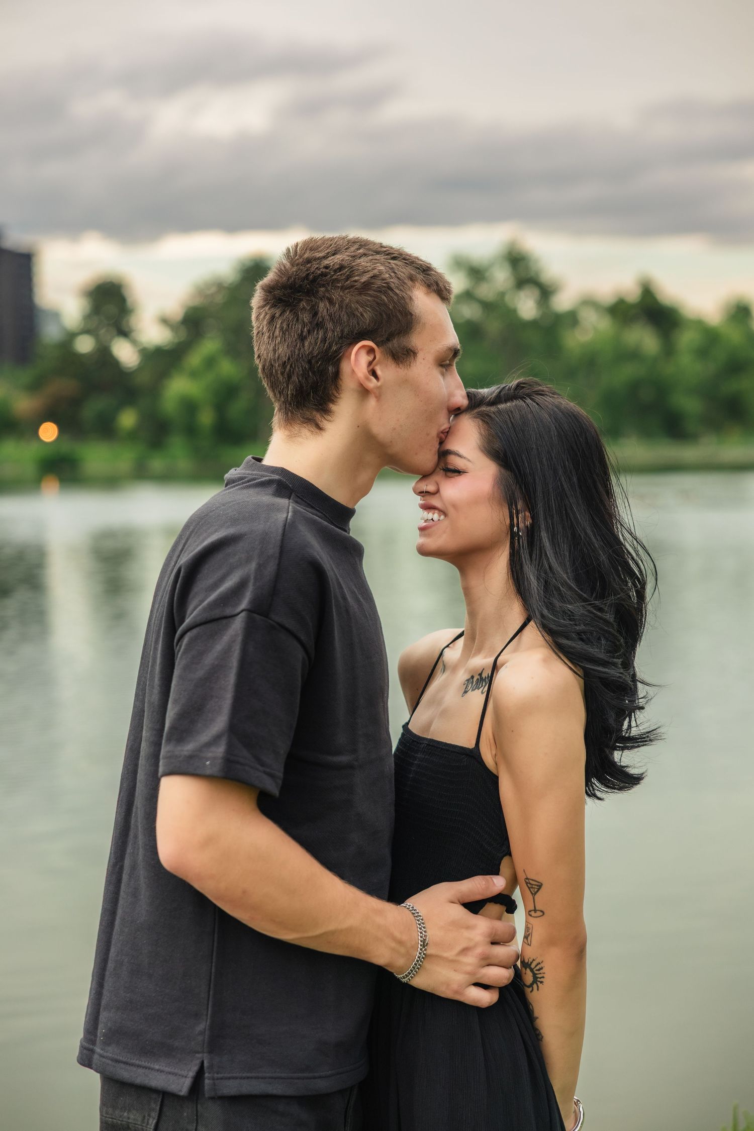 A young couple shares a tender moment embracing and kissing near a serene lake with lush green trees in the background.