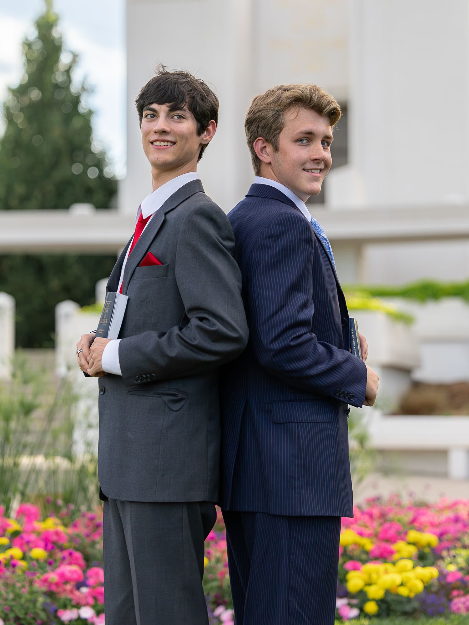 Two young men in suits pose back-to-back outdoors near colorful flowers, smiling for a formal portrait.