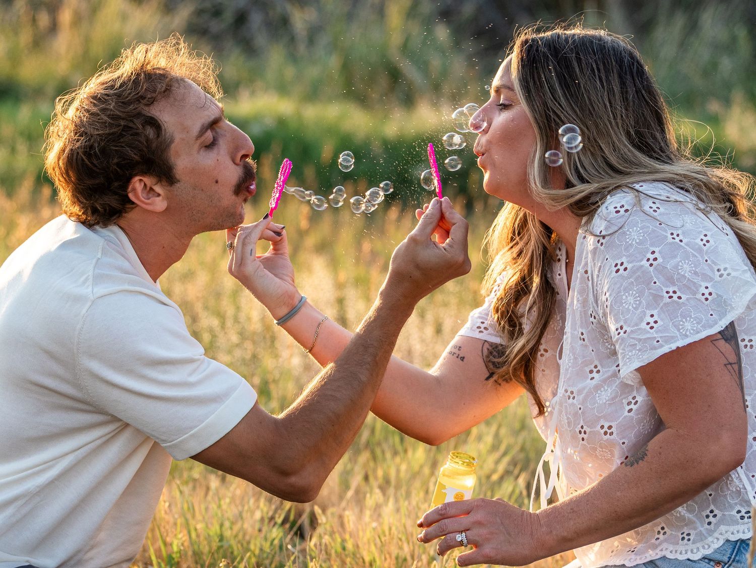 Couple blowing bubbles together outdoors in a sunny field, sharing a playful and romantic moment.