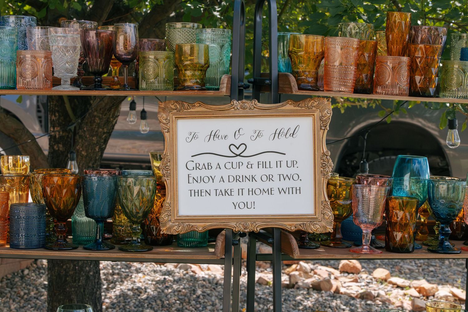 Colorful vintage glassware display at wedding bar with sign inviting guests to grab a cup and take it home.