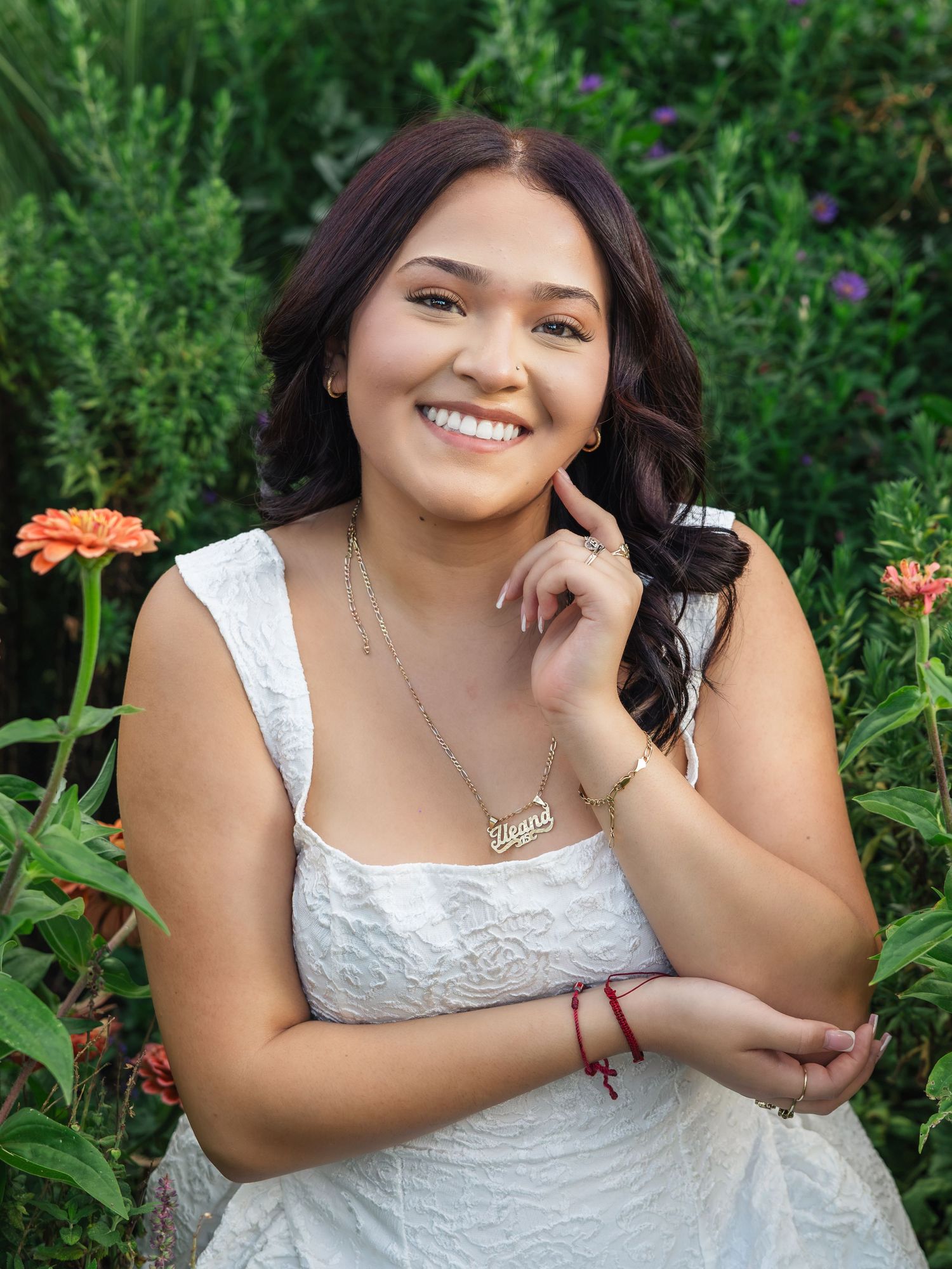 Young woman in white dress smiling among orange flowers and green foliage, wearing a name necklace and red bracelet.