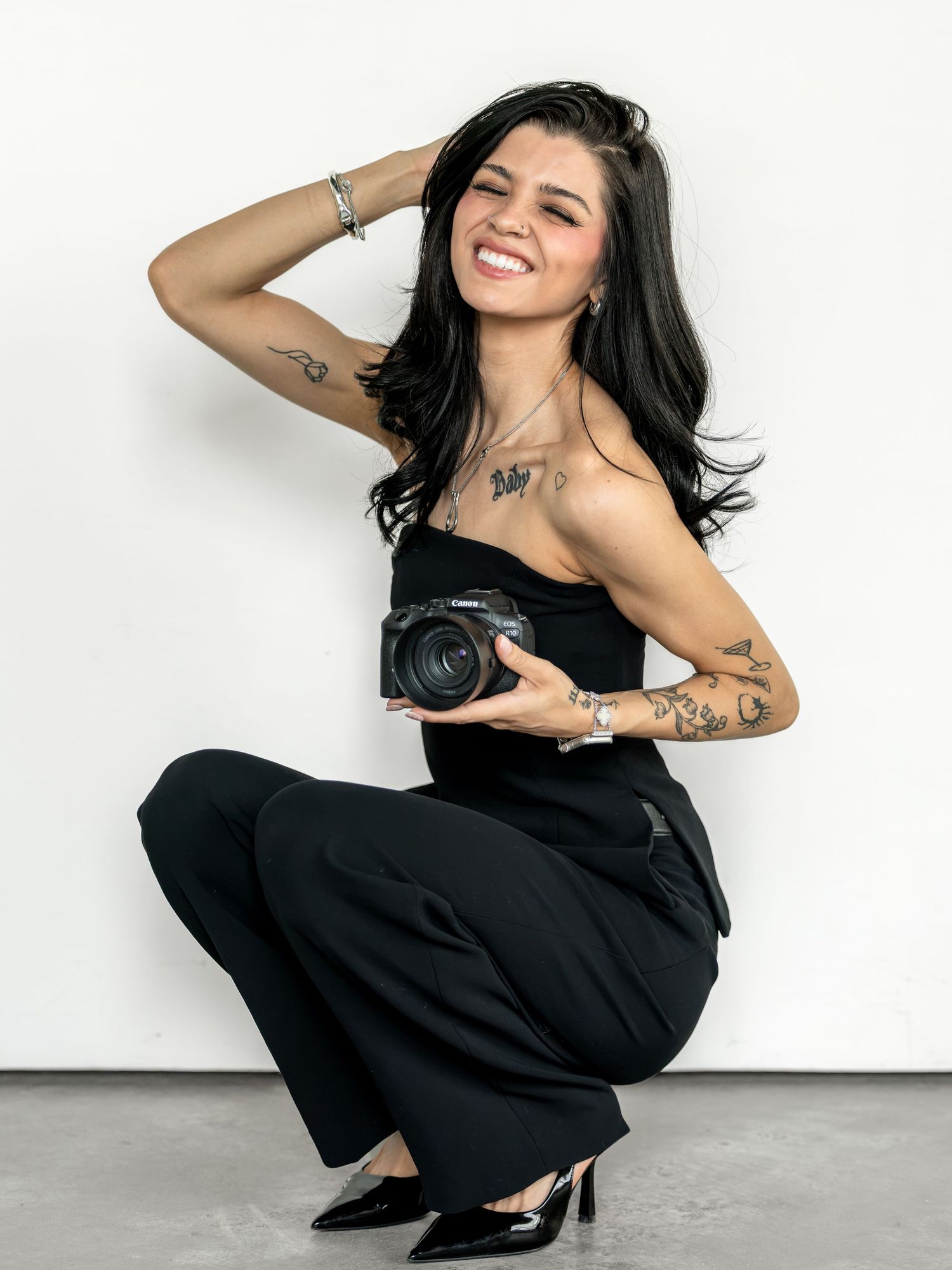 Smiling woman in black outfit crouching while holding a camera, with tattoos and long dark hair.