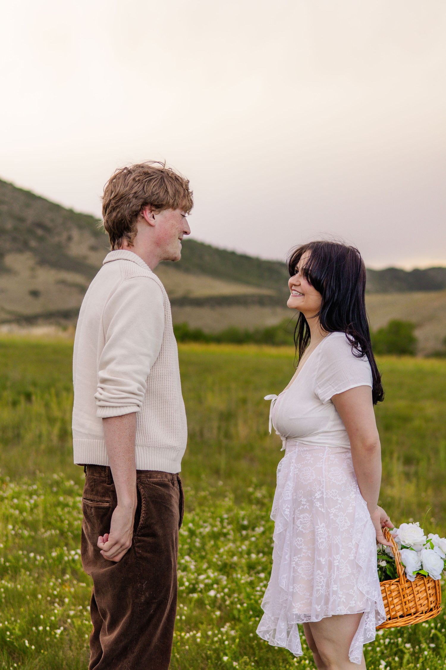A young couple smiles at each other in a green meadow, woman holding a wicker basket with white flowers.