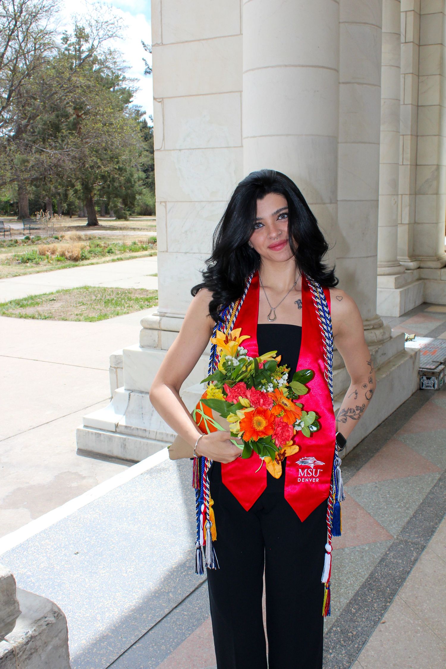 Woman in black outfit wearing MSU Denver graduation stoles holding colorful flower bouquet outdoors.