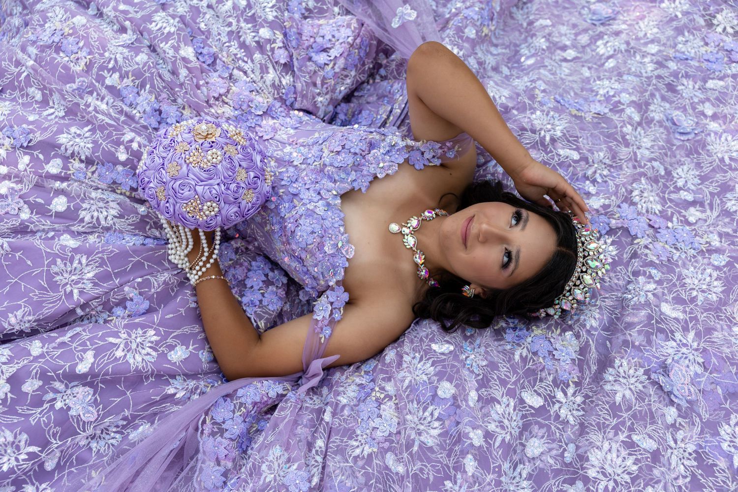 Young woman in lavish purple floral quinceañera gown lying gracefully, adorned with pearl jewelry and floral crown.