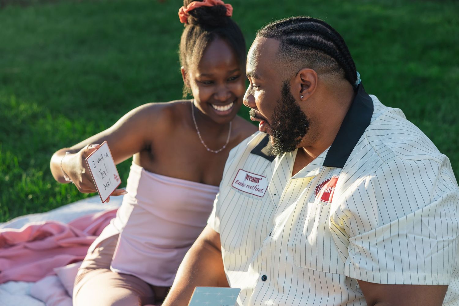 A joyful couple laughing together outdoors on a pink blanket, sharing a card during a sunny picnic.
