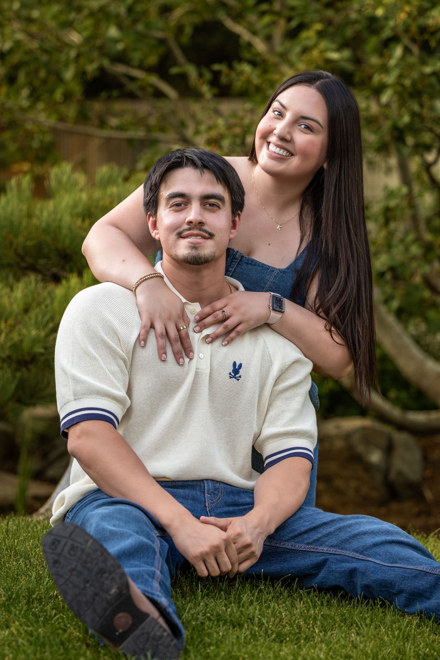Young couple posing outdoors on grass, woman smiling behind seated man in white polo, green garden background.