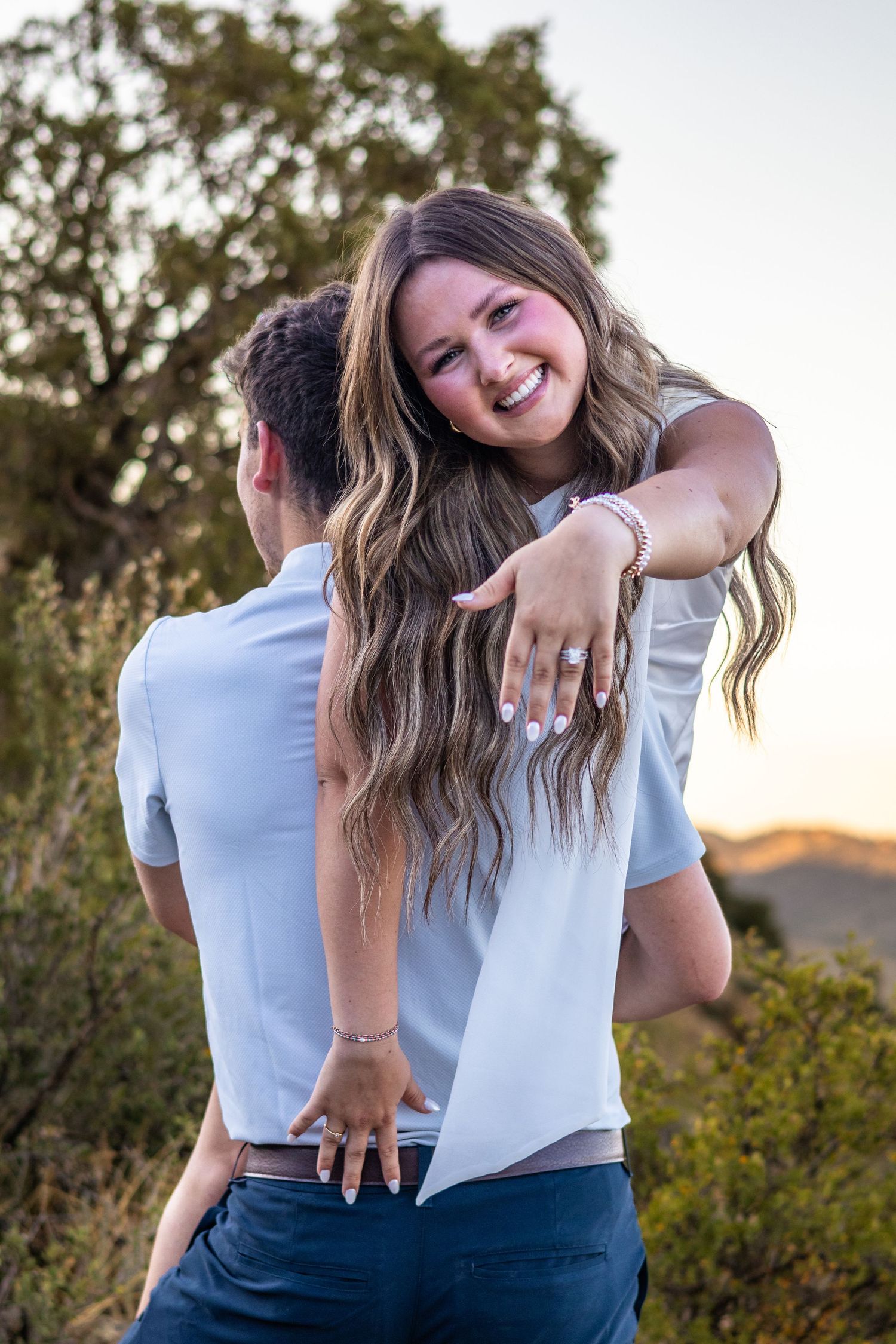 Smiling woman with long wavy hair hugs man outdoors at sunset, showing off engagement ring on her hand.