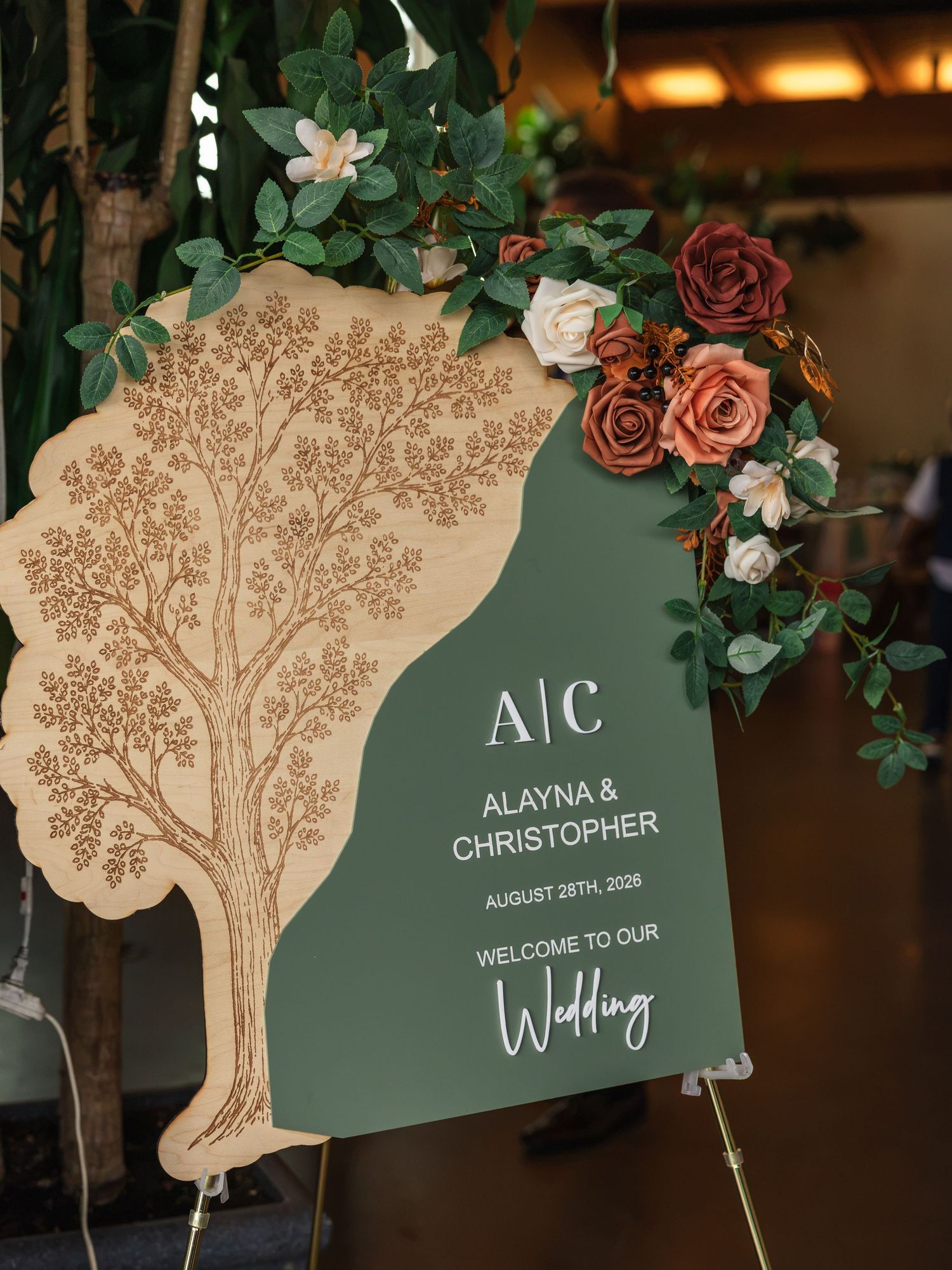 Elegant wedding welcome sign with tree engraving, green panel, floral arrangement, reading Alayna & Christopher August 28th 2026.