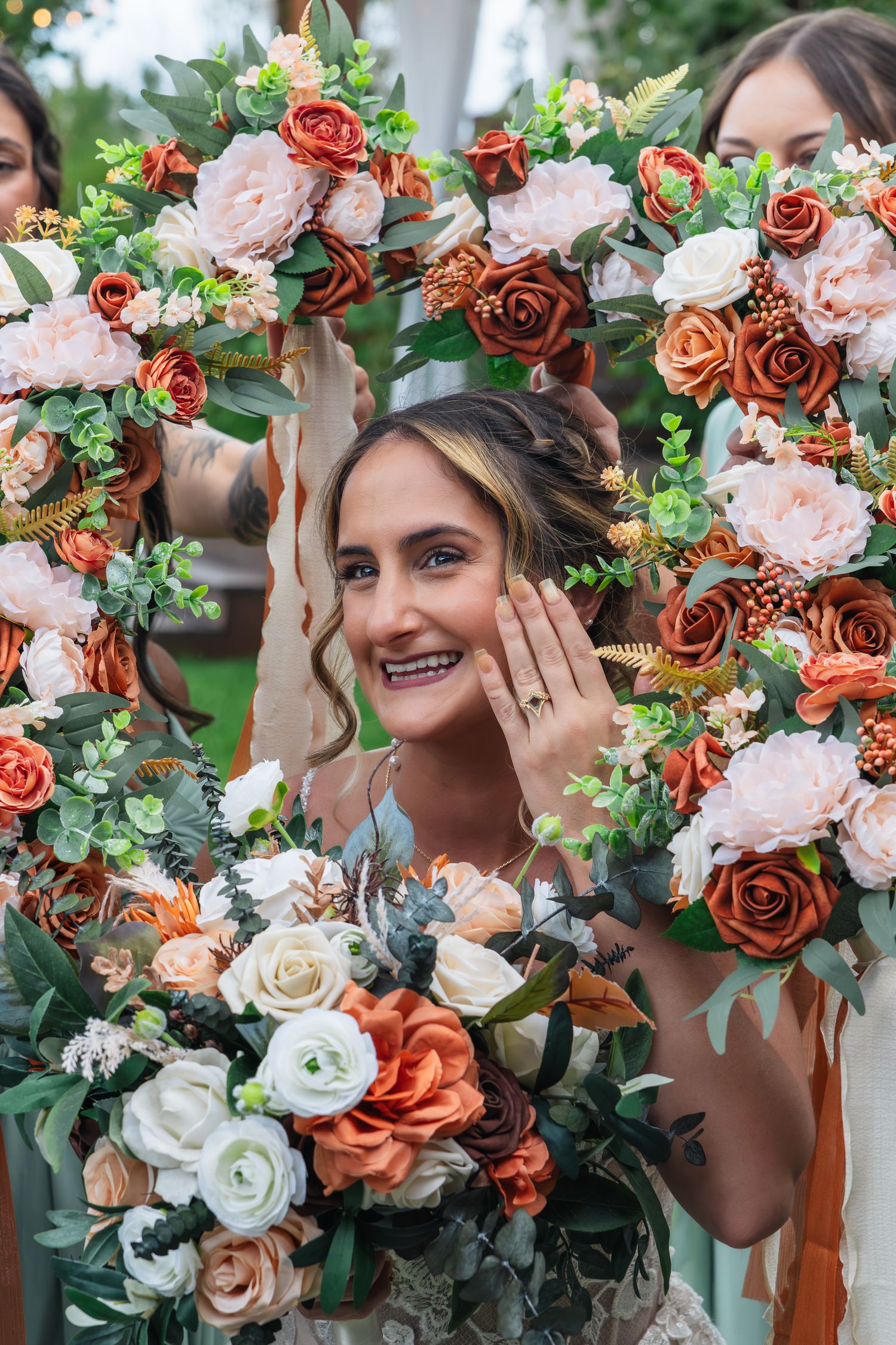 Smiling bride shows off engagement ring framed by floral wreaths with rust and blush roses at wedding.