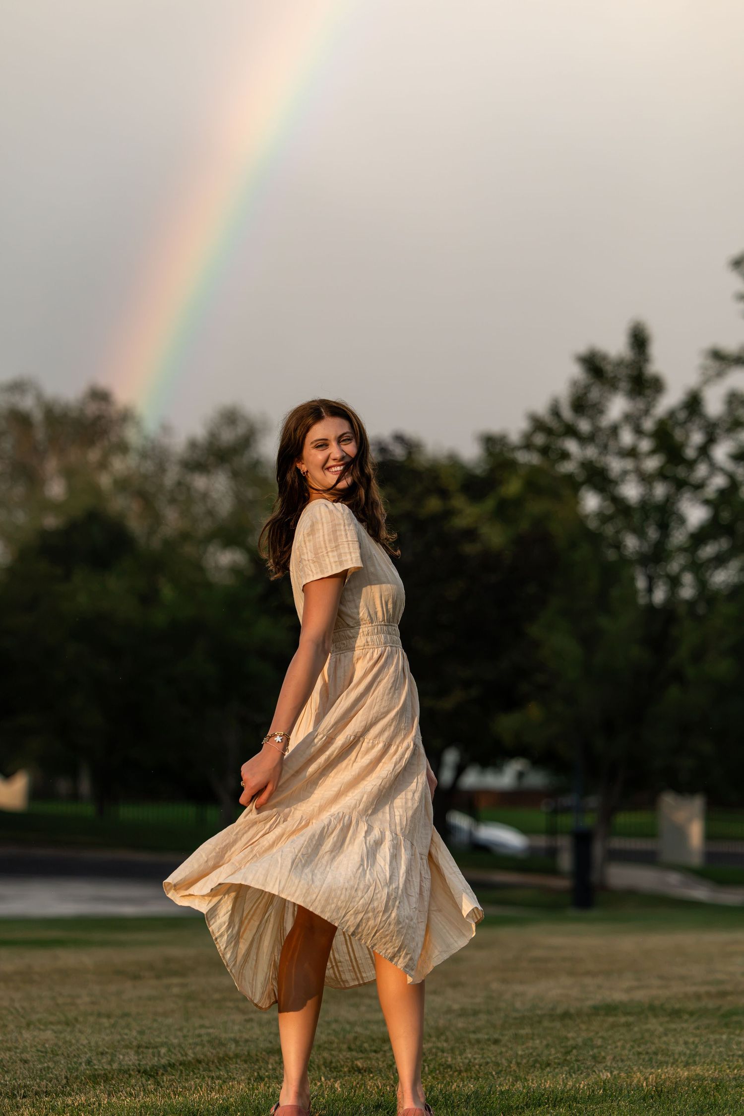 Young woman in flowing beige dress twirling in park with vibrant rainbow in sky behind her.