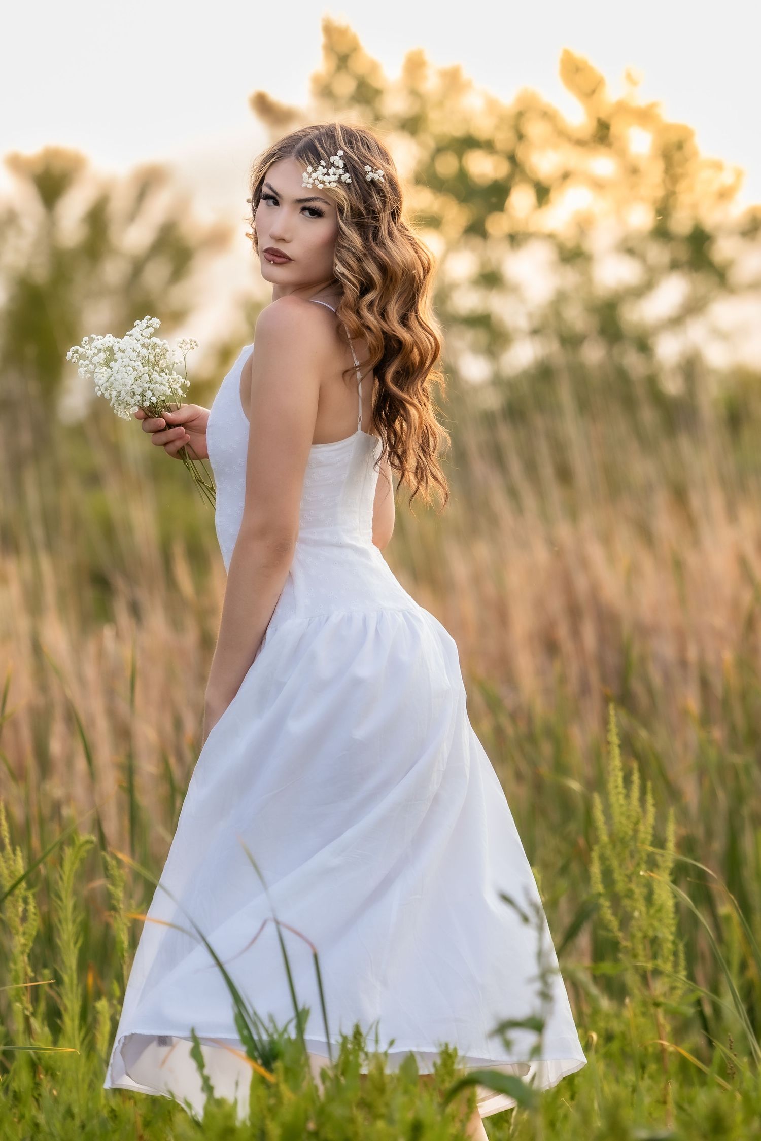 Young bride in white dress holding wildflowers in a sunlit field, with wavy hair and floral crown.