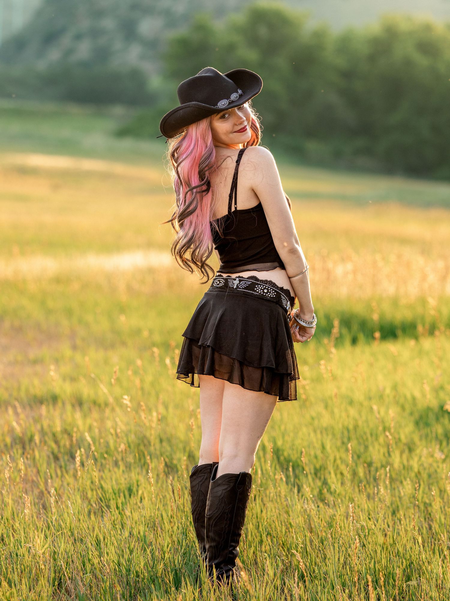Young woman in black cowboy hat, mini skirt, and boots posing in a sunny green field.