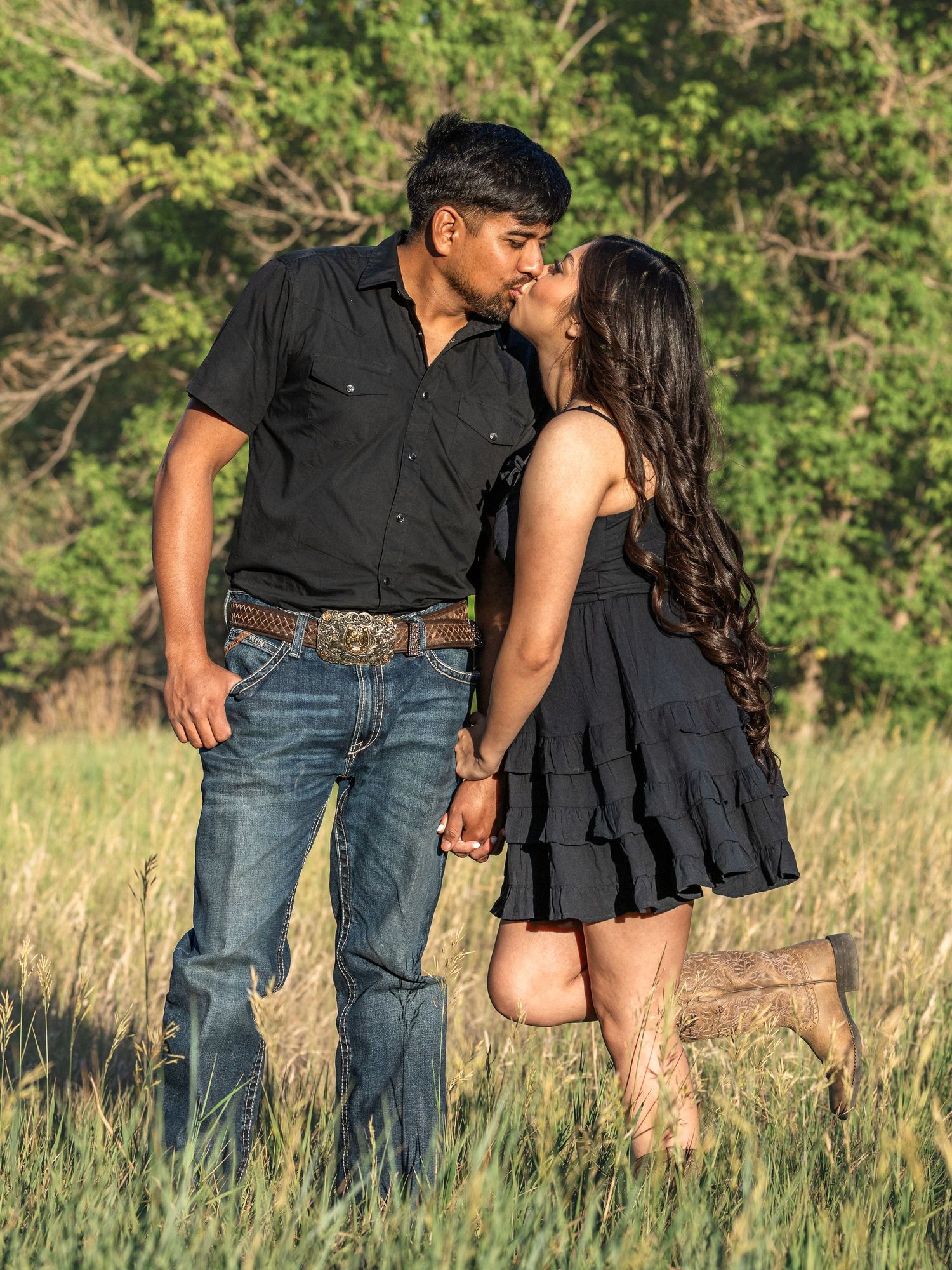 Couple sharing a kiss in a sunlit field, wearing black outfits and boots, surrounded by green trees.
