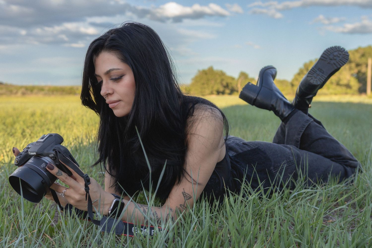 Woman with black hair lying in grass, reviewing photos on a DSLR camera in an open field under a cloudy sky.