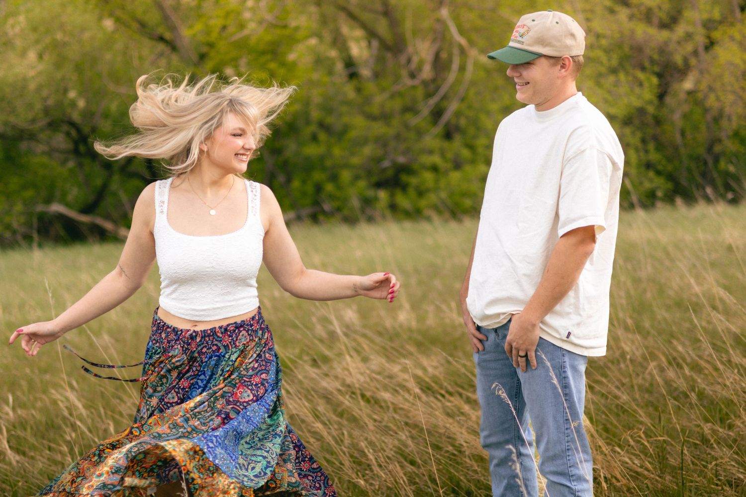Blonde woman in white crop top and colorful skirt spinning joyfully while man in cap watches in grassy field.