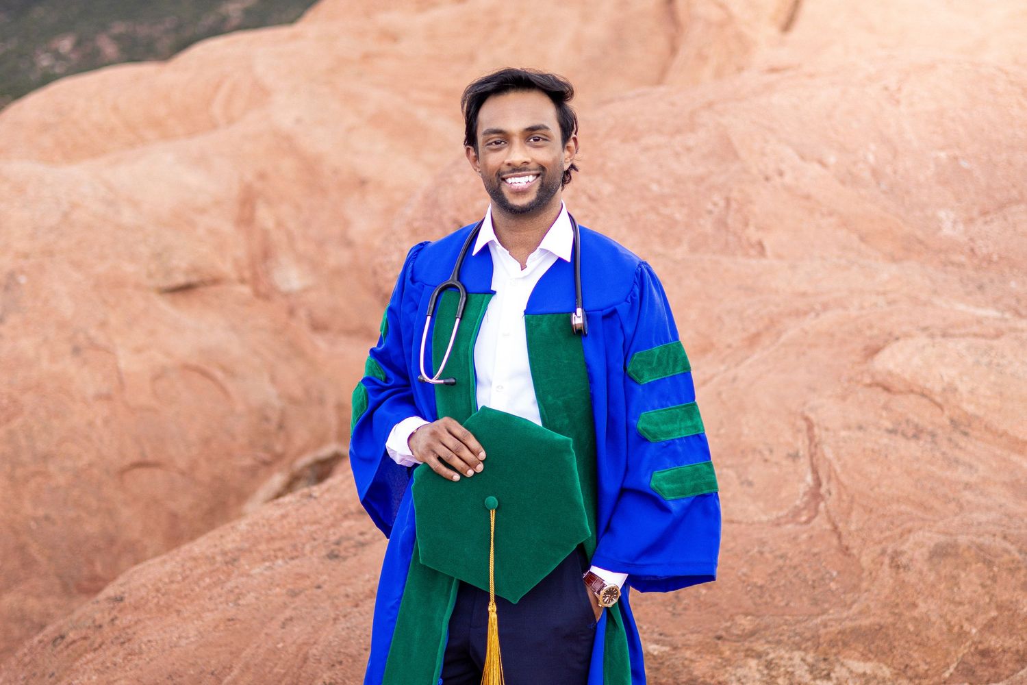 Smiling medical graduate in blue doctoral robes with stethoscope holding green cap at red rock formation.