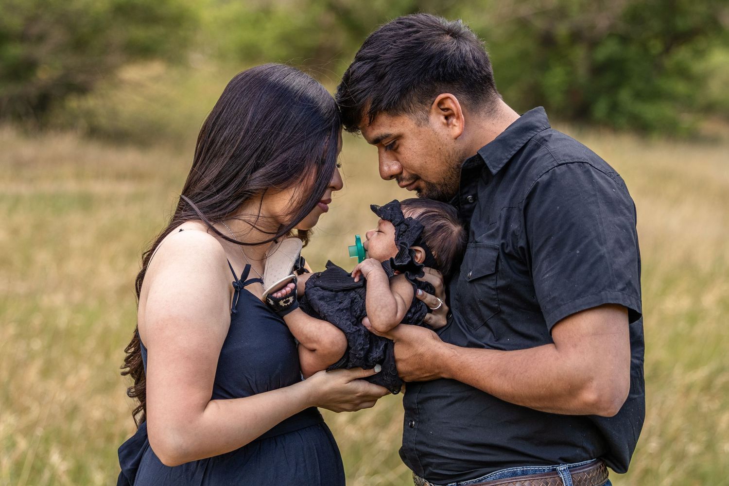 A couple tenderly holds their newborn baby outdoors in a green field during a family portrait session.