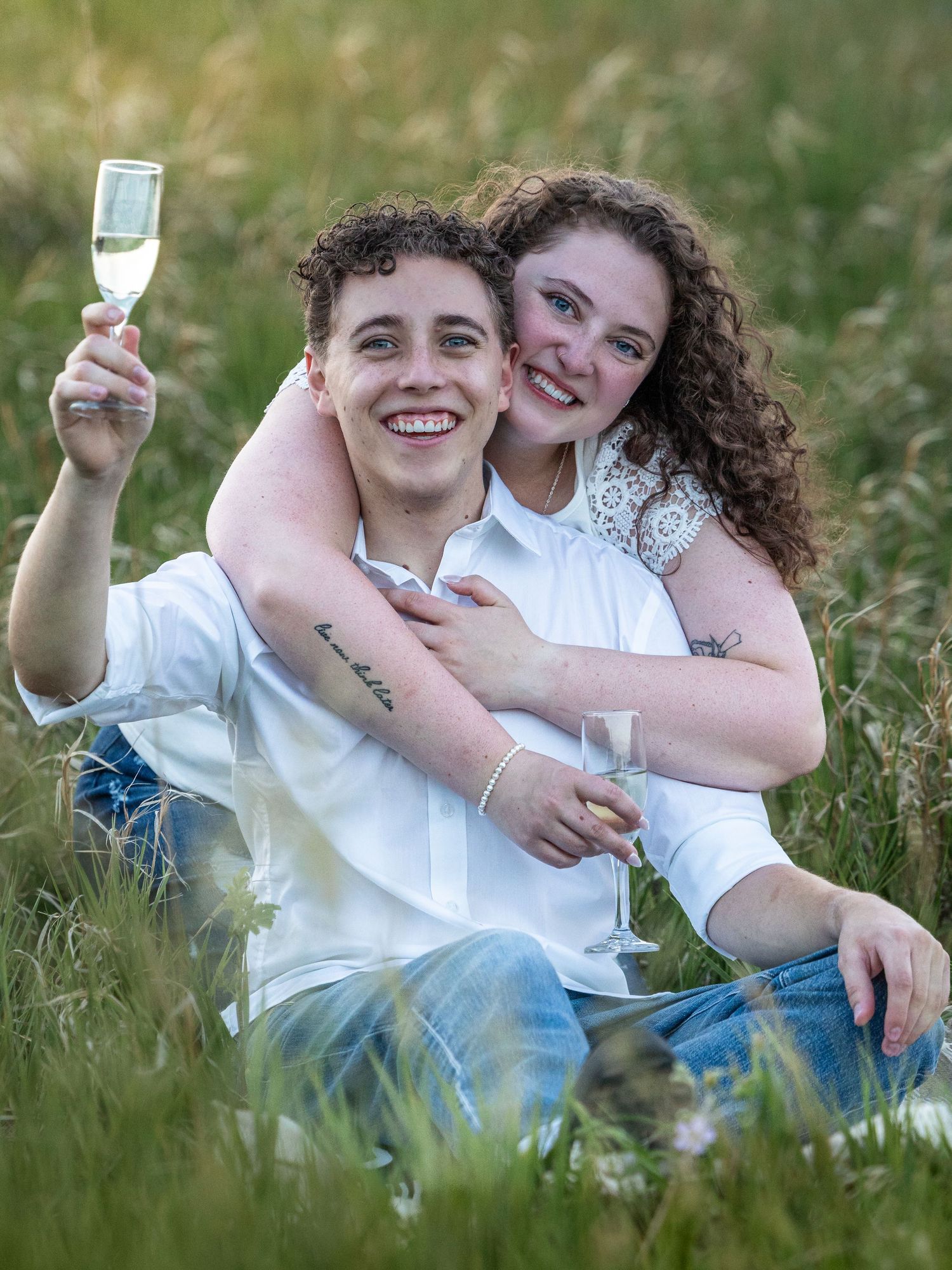 Happy young couple celebrating in a meadow, holding champagne glasses, wearing white shirts, smiling outdoors.