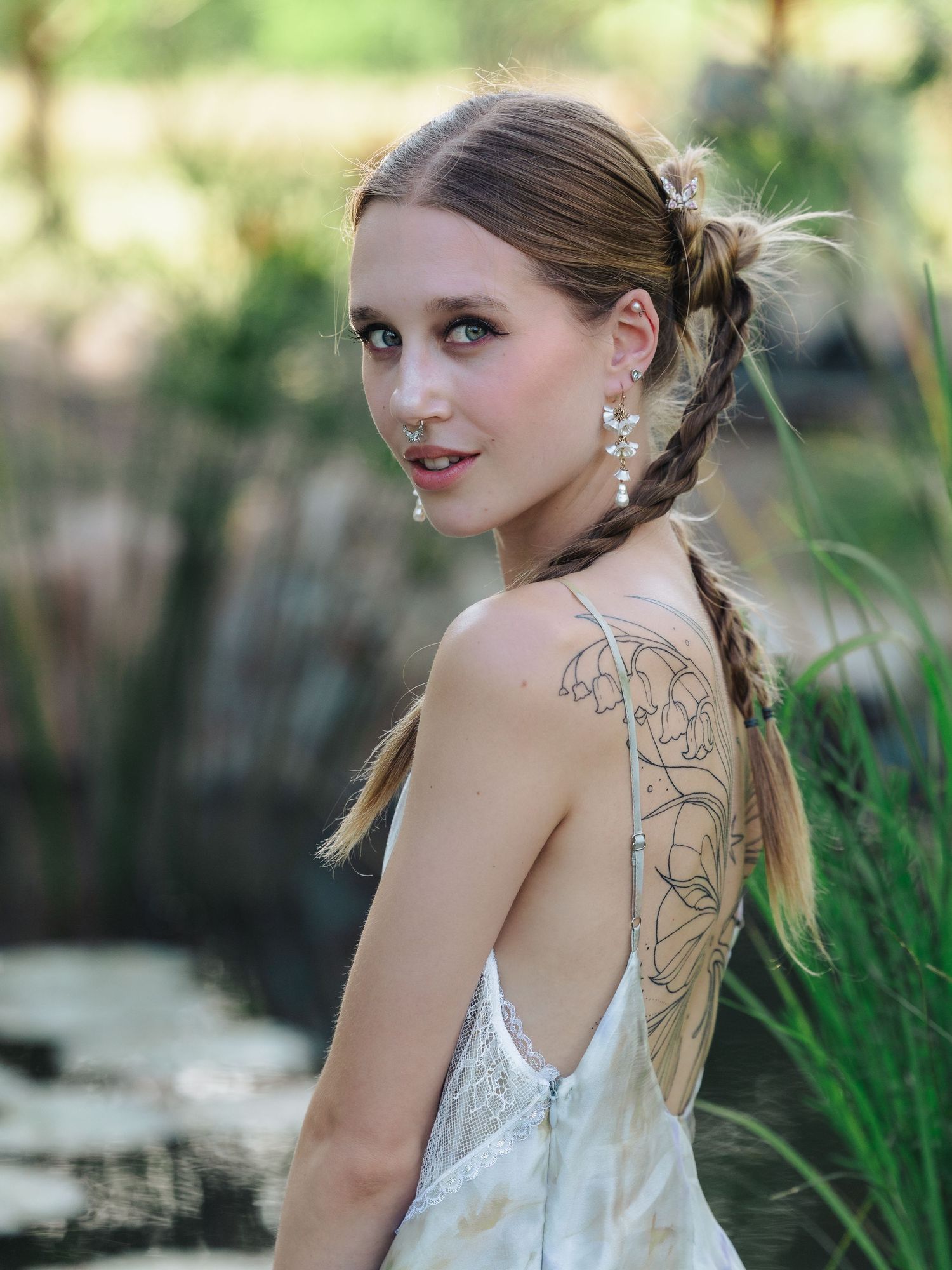 Young woman in backless wedding dress with floral tattoo, braided hair, and pearl earrings in a garden setting.