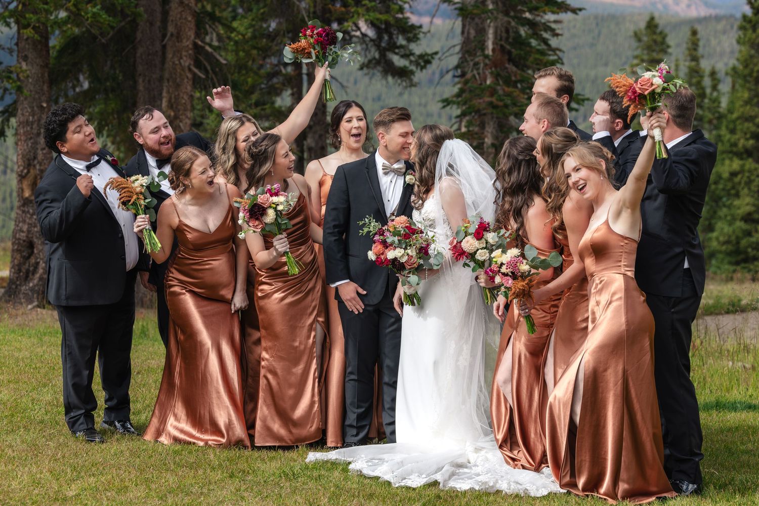 Wedding party in bronze gowns and tuxedos celebrate outdoors with bouquets, bride and groom kissing in center.