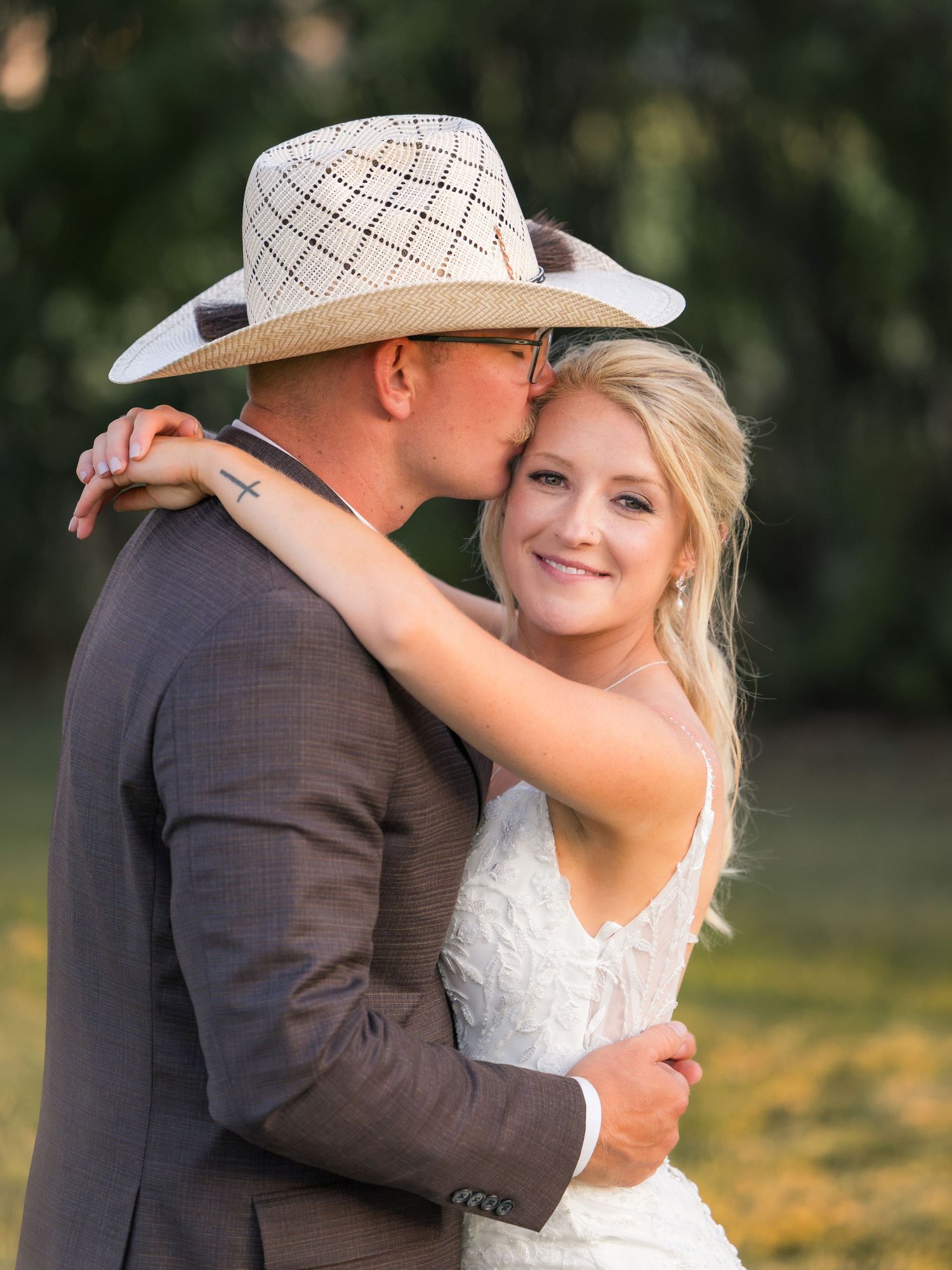 Bride and groom embrace outdoors, groom wearing cowboy hat kissing bride on temple, both dressed in wedding attire.