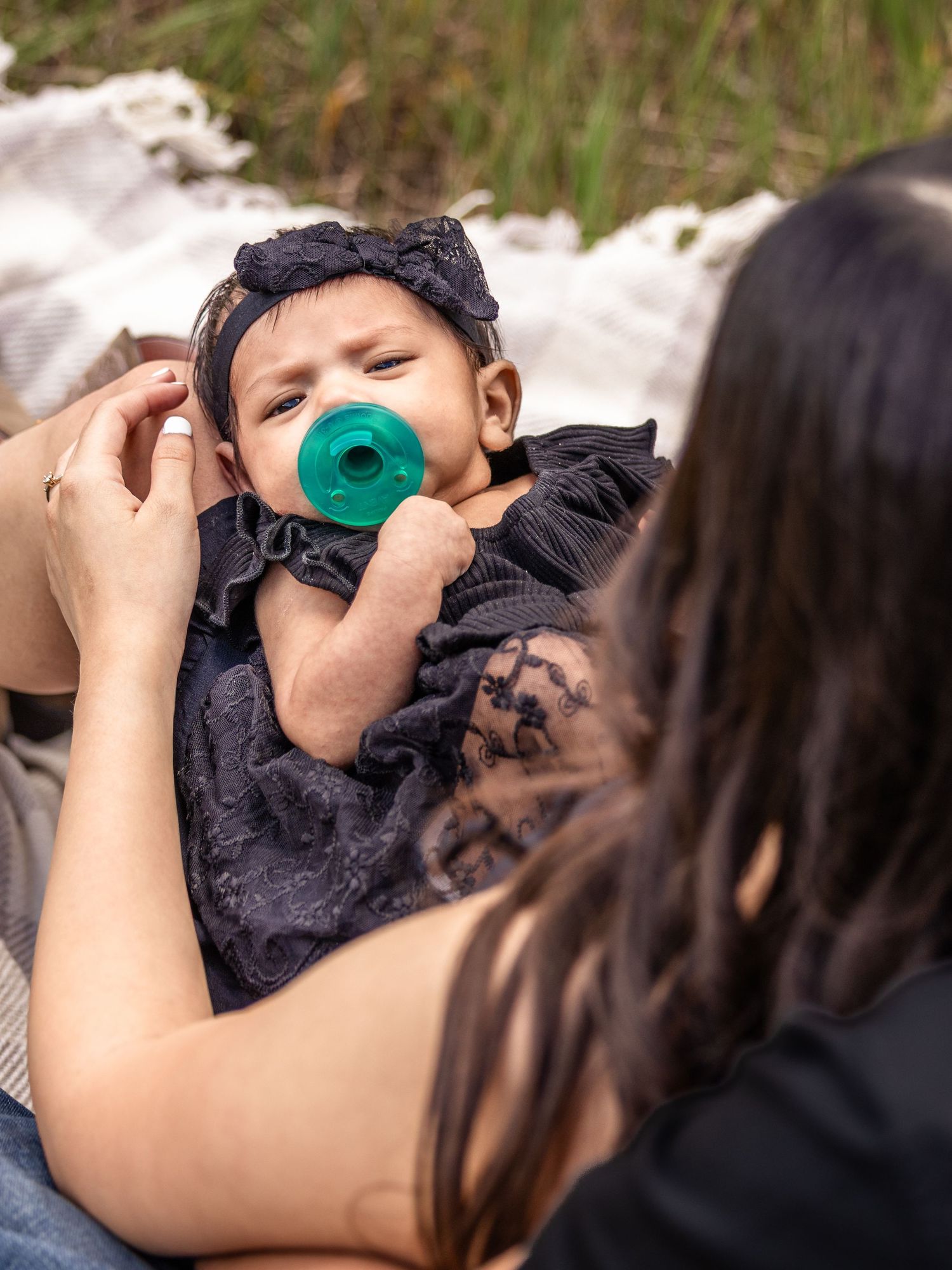 A mother gazes lovingly at her baby girl in a black dress with a teal pacifier, lying on a blanket outdoors.