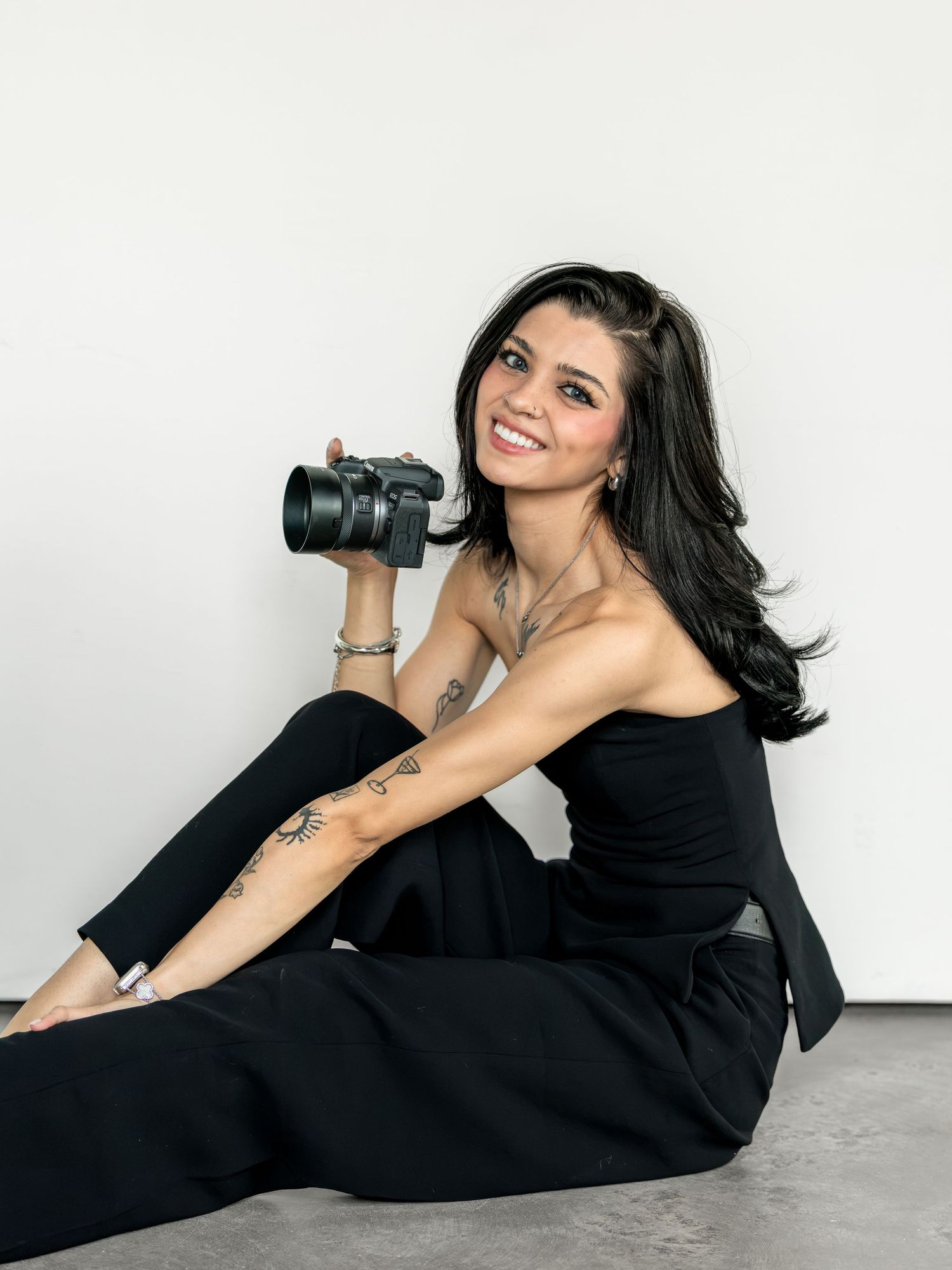 Woman in black outfit sitting on floor holding a camera, smiling, with tattoos visible on her arm.