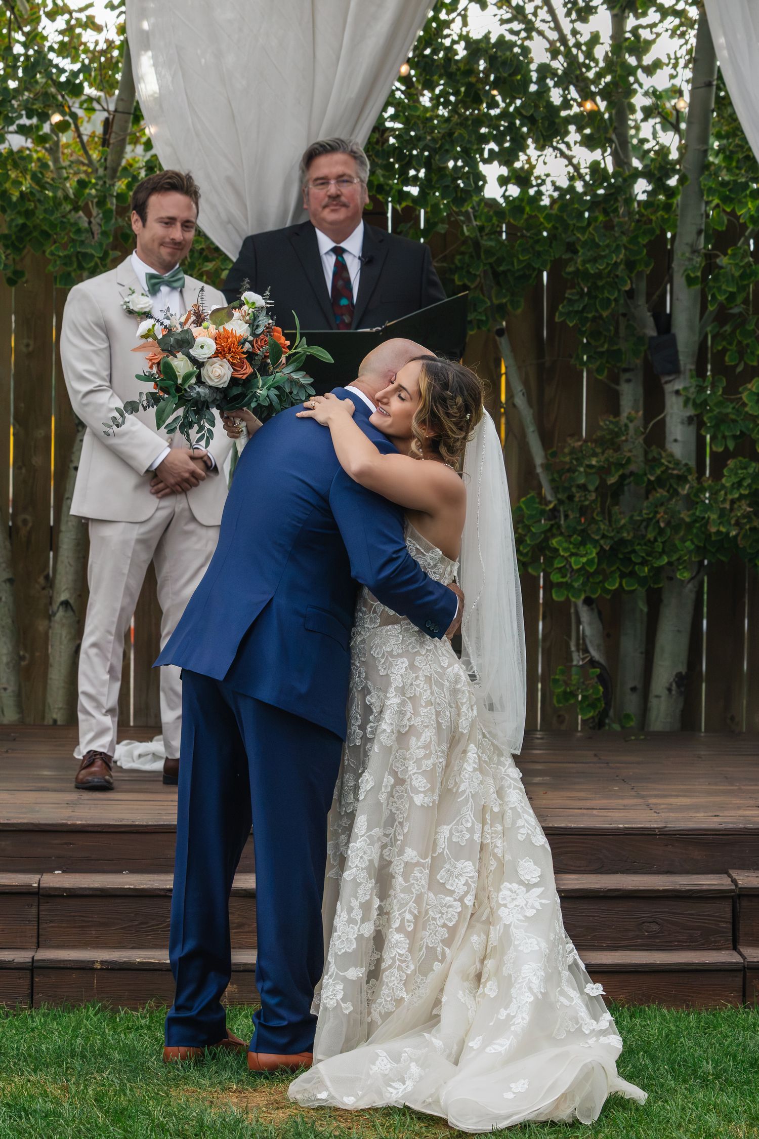 Bride and groom share first kiss at outdoor wedding ceremony with officiant and groomsman watching.