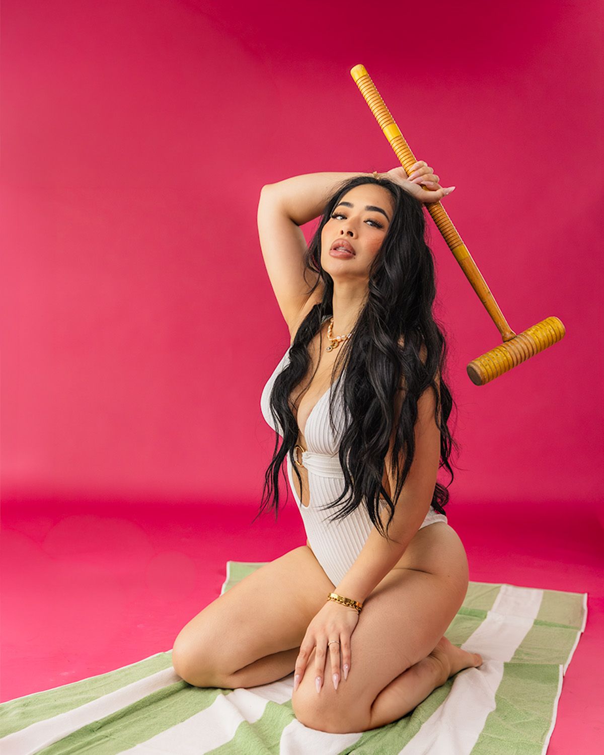 Woman in white swimsuit poses on striped towel holding a croquet mallet against a vibrant pink background.