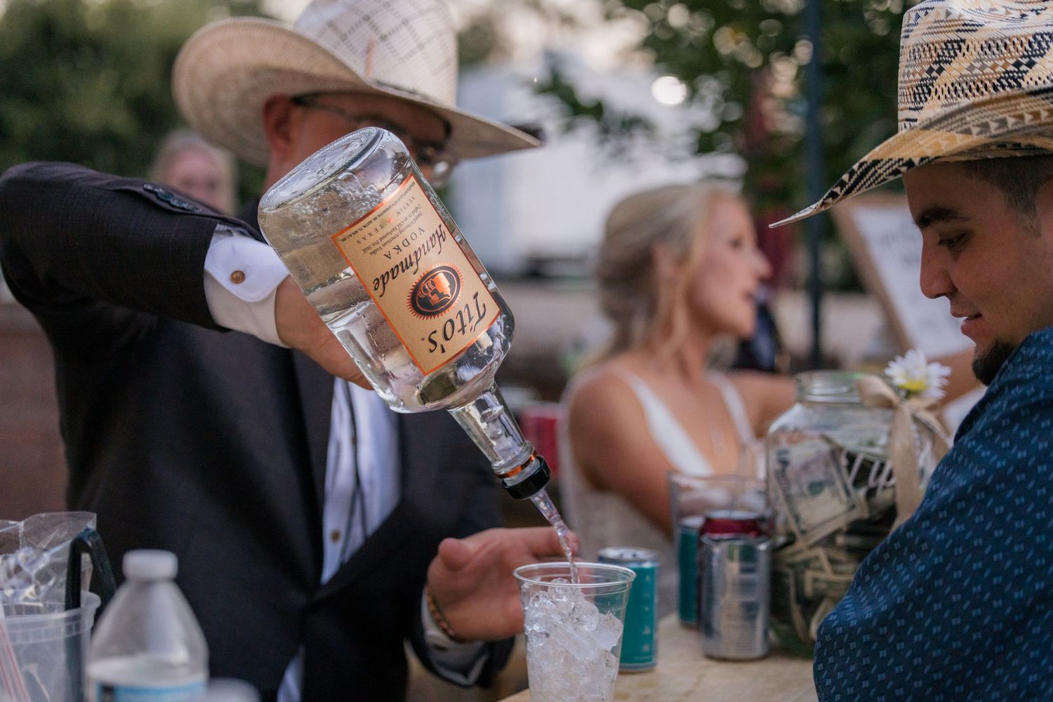 Guest in cowboy hat pours large bottle of Tito's vodka at outdoor rustic wedding reception table.