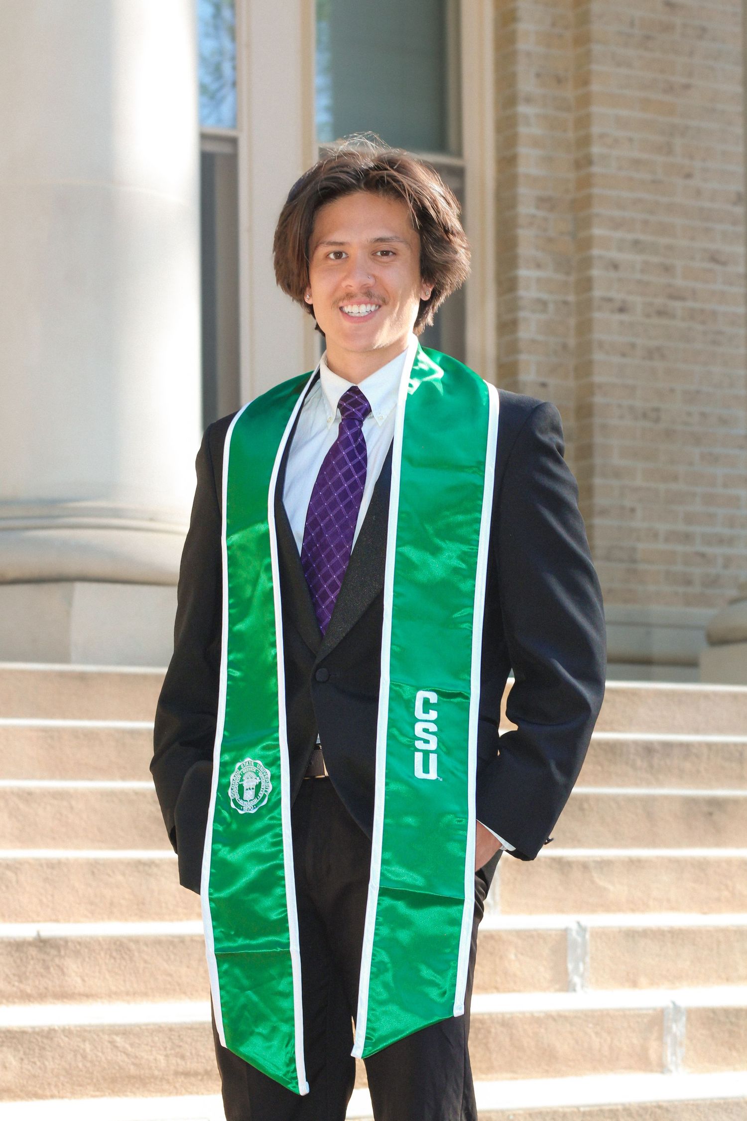 Young man in black suit with green graduation stole smiling on steps at outdoor graduation ceremony.