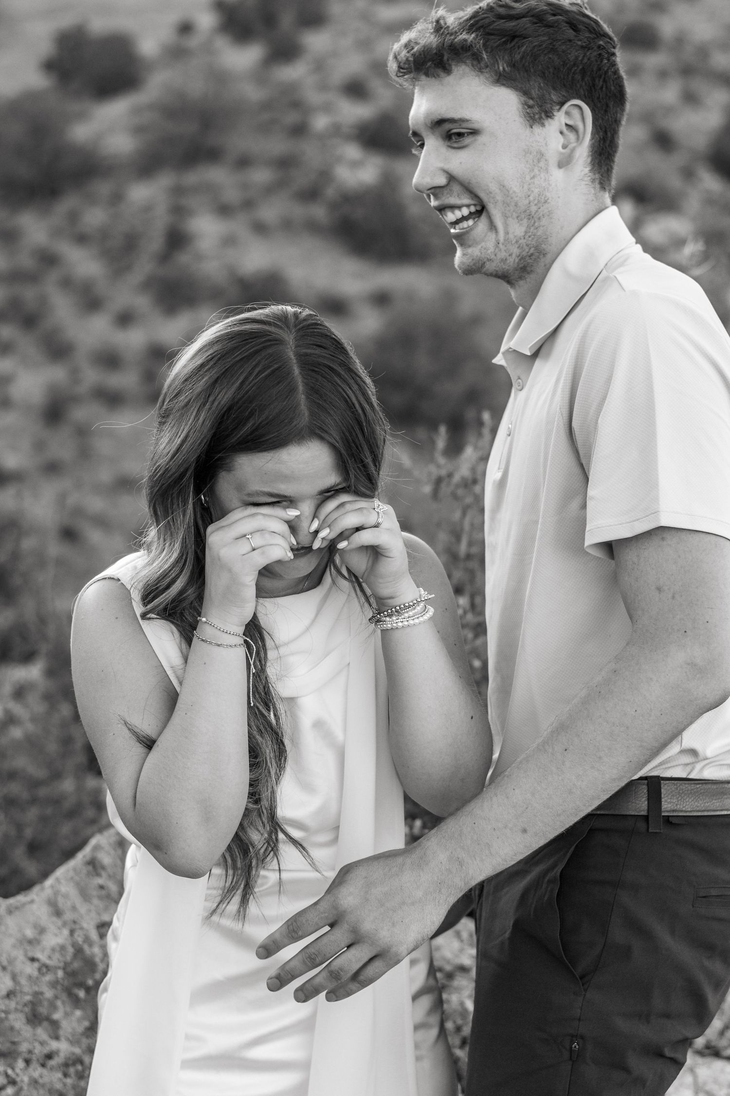 Black and white photo of a laughing couple outdoors, woman covering her face with hands, man smiling beside her.