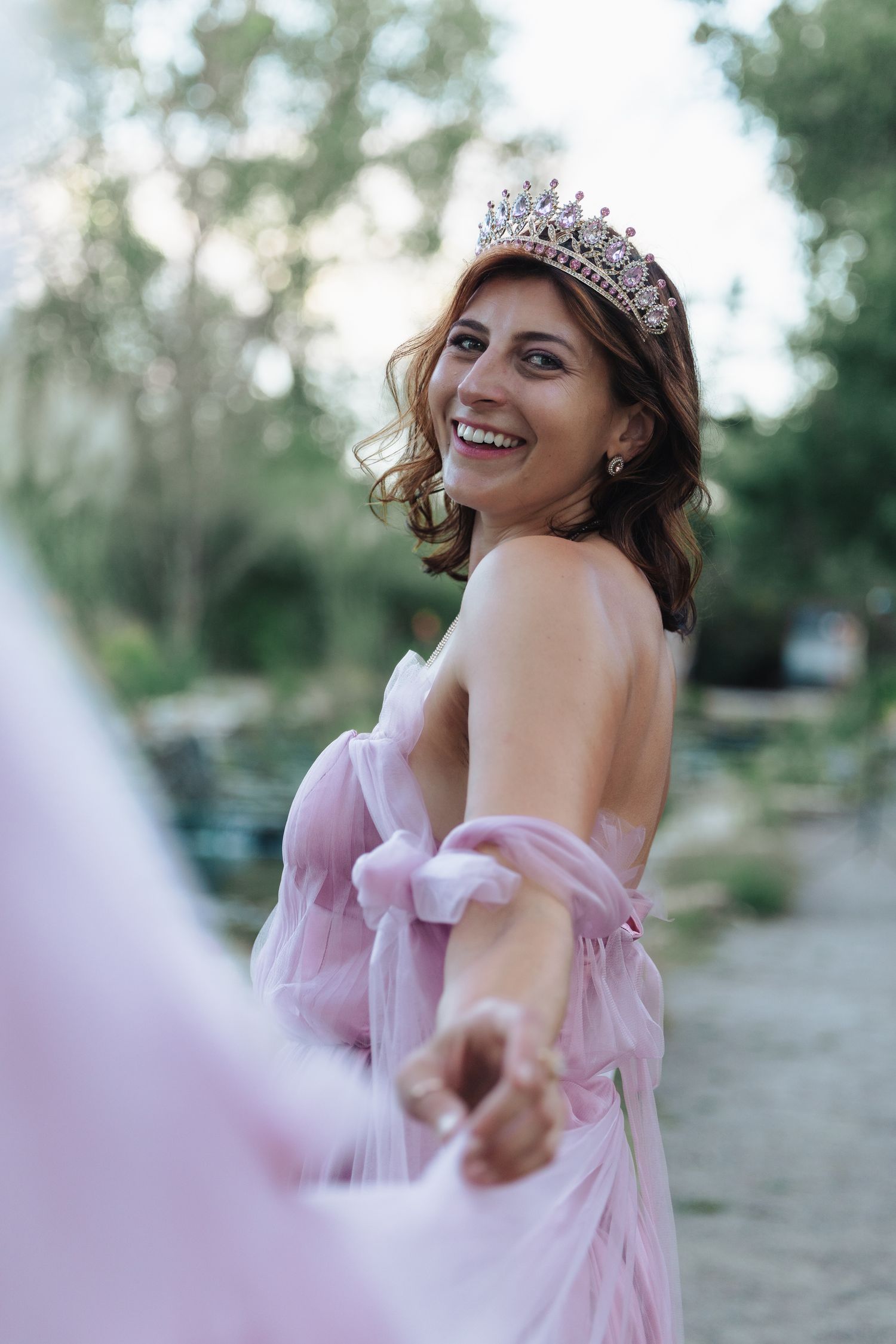 Smiling bride in pink tulle gown and diamond tiara holds hands outdoors in lush garden setting.