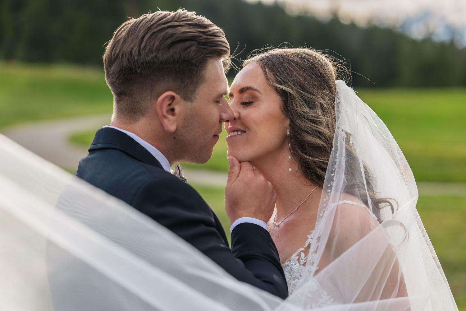 A bride and groom share a romantic moment outdoors, with her veil flowing around them against a lush green mountain backdrop.
