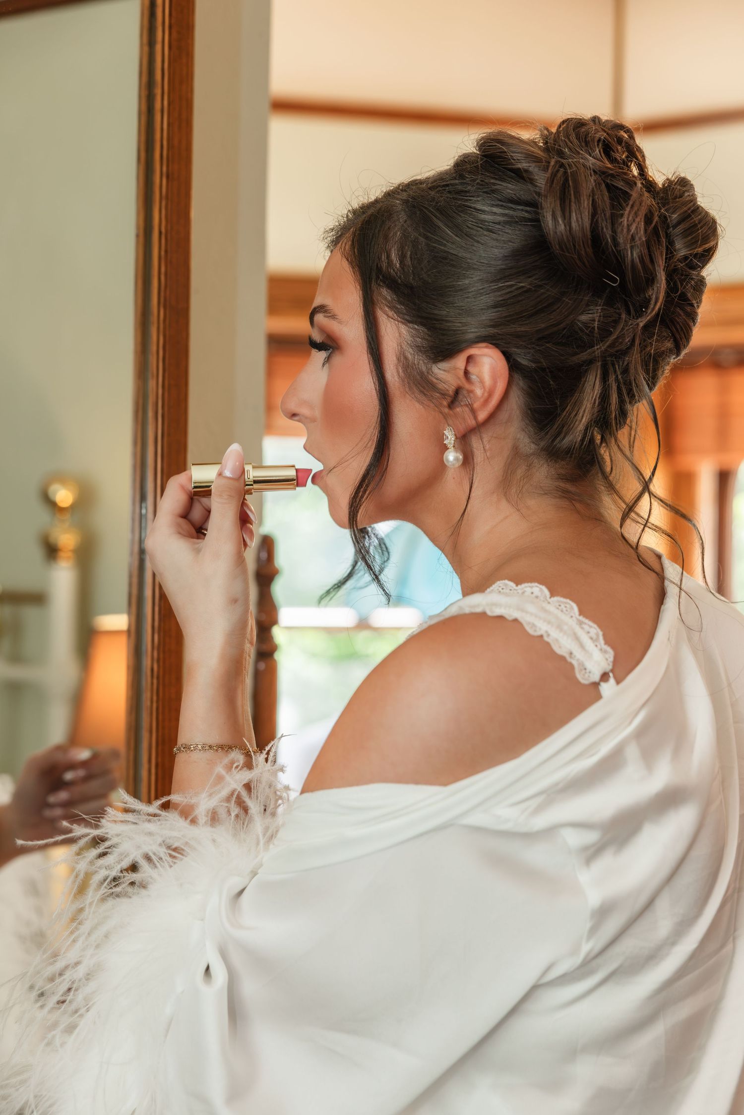 Bride in white feather robe applying red lipstick while looking in mirror, elegant updo and drop earrings.